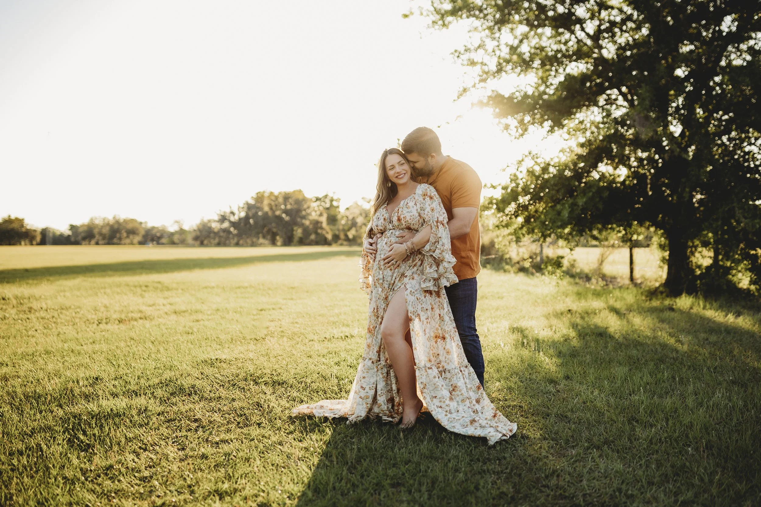 Romantic maternity photo of an expectant couple in a golden field at sunset, sharing a quiet moment as the mother holds her baby bump and her partner embraces her from behind.
