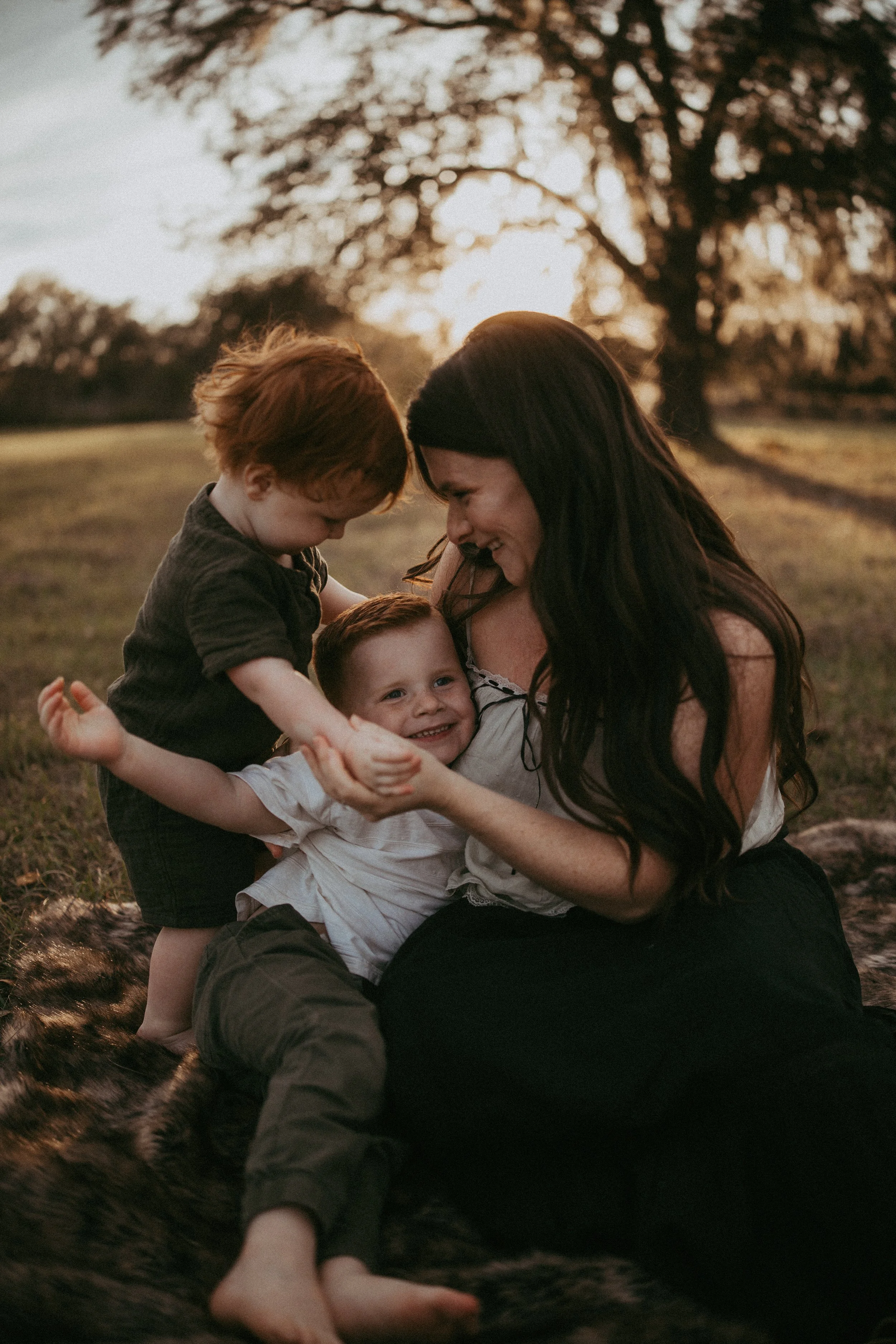 A mother laughs with her young sons as they cuddle together on a blanket in a grassy field.