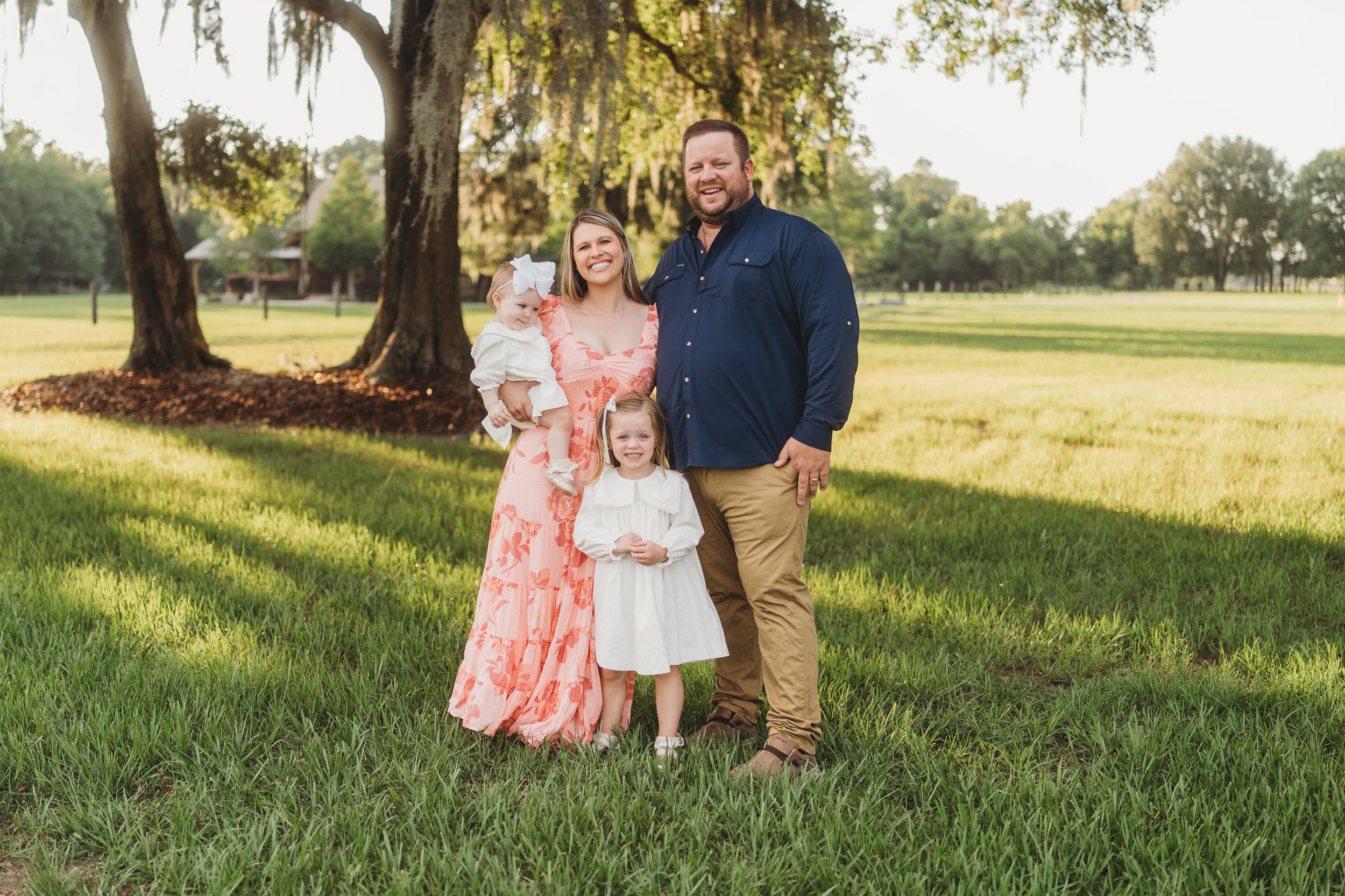 A smiling family of four stands together beneath majestic oak trees during a golden-hour portrait, with parents holding their young daughters in a peaceful outdoor setting that celebrates love, connection, and the beauty of everyday family life.