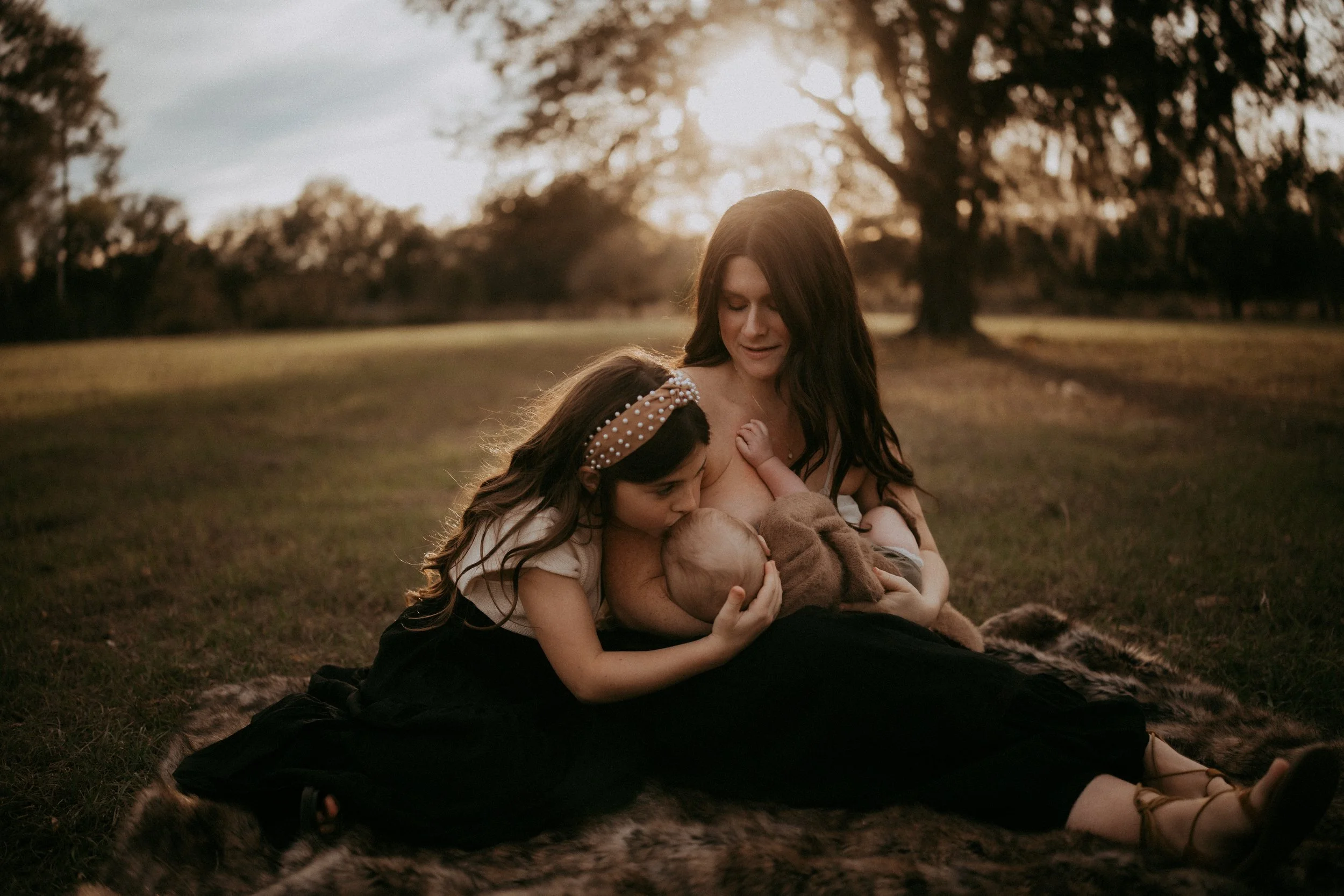 Mother tandem nursing her baby and older daughter while sitting together in a sunlit field during a family photography session in Northeast Florida.