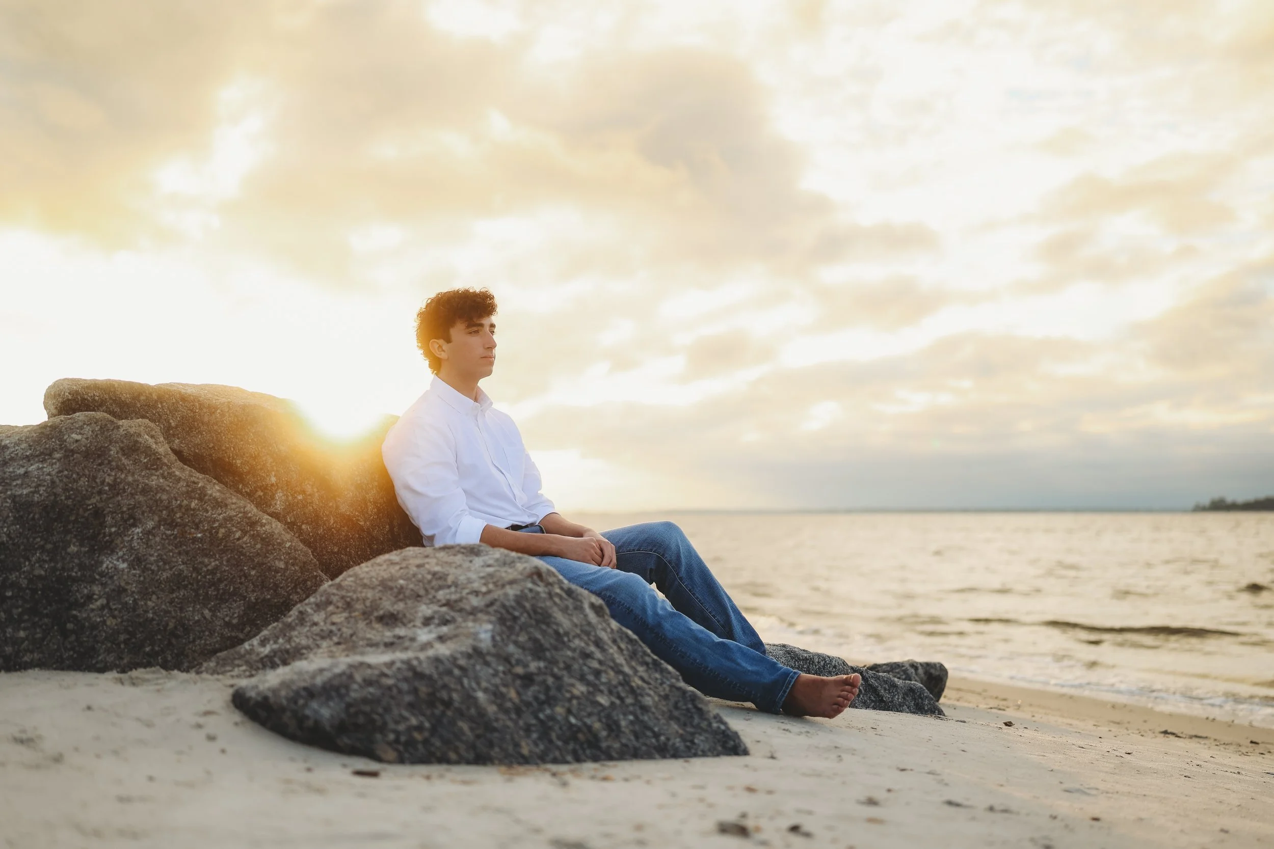 A teenage son sits quietly on the beach rocks as the sun sets over the water behind him.