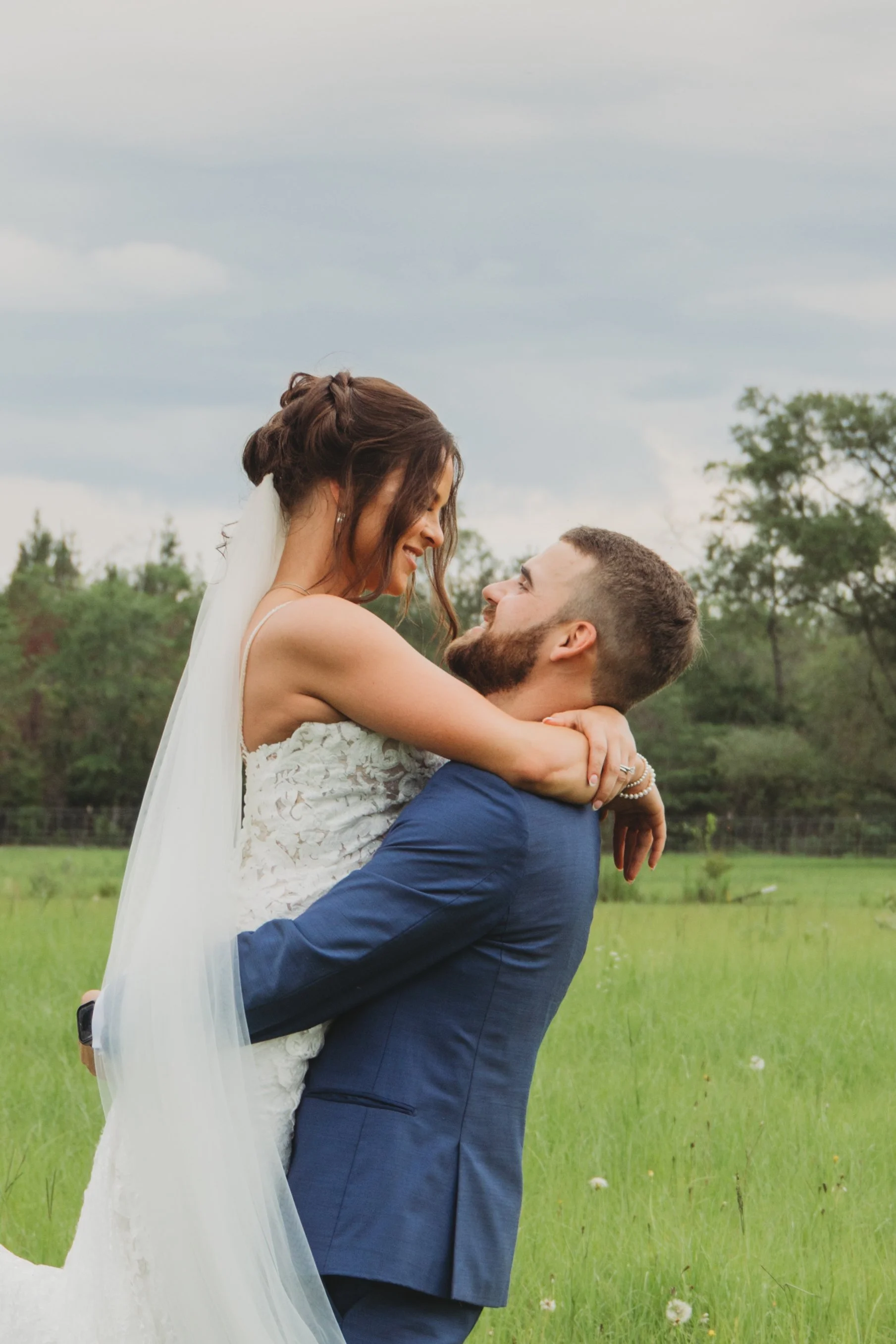 Dylan lifts Emma as they smile at each other in a green field beneath the cloudy sky at Red River Estate.