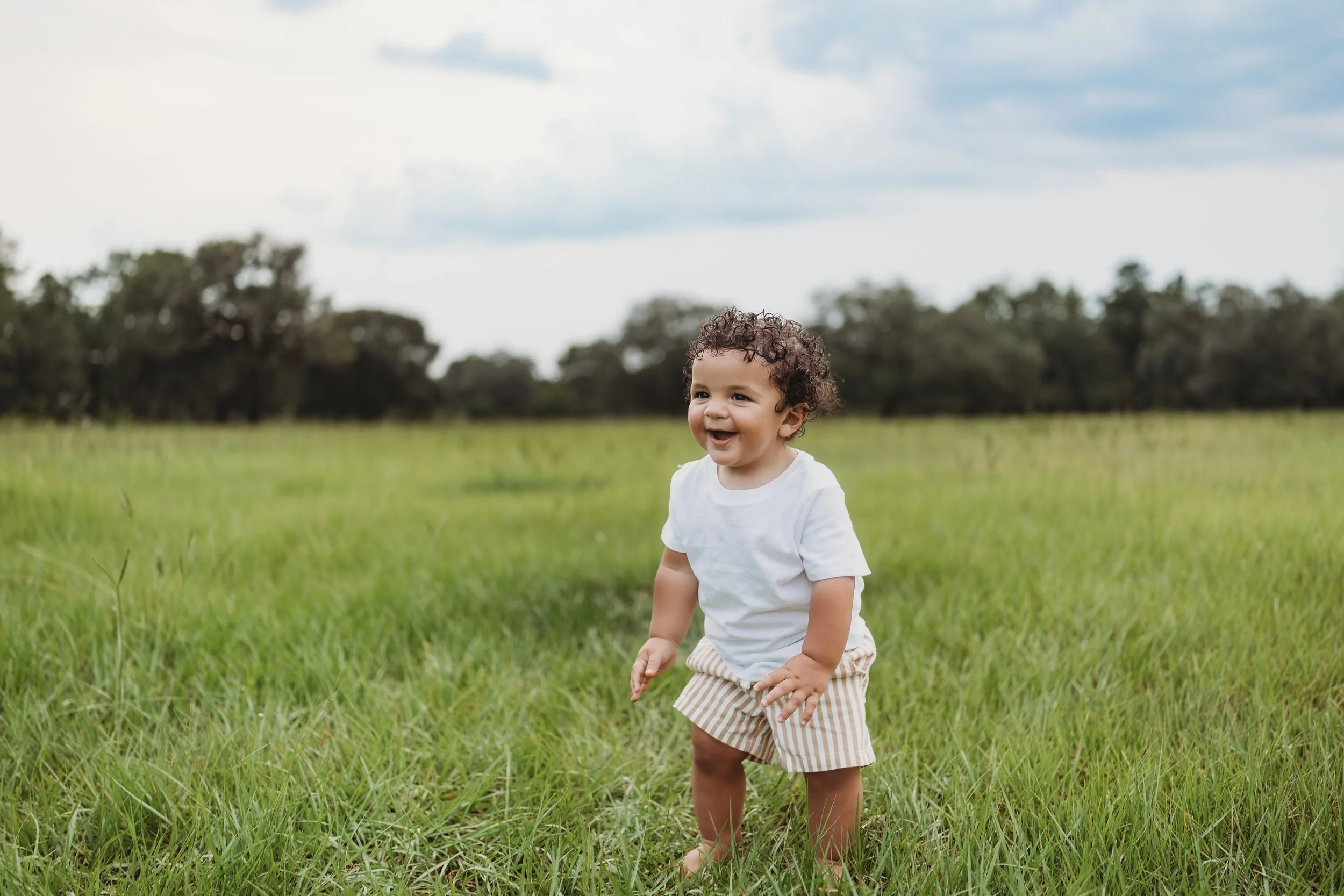 Toddler laughing while standing barefoot in an open grassy field during a relaxed outdoor portrait session, capturing the simple joy of childhood.
