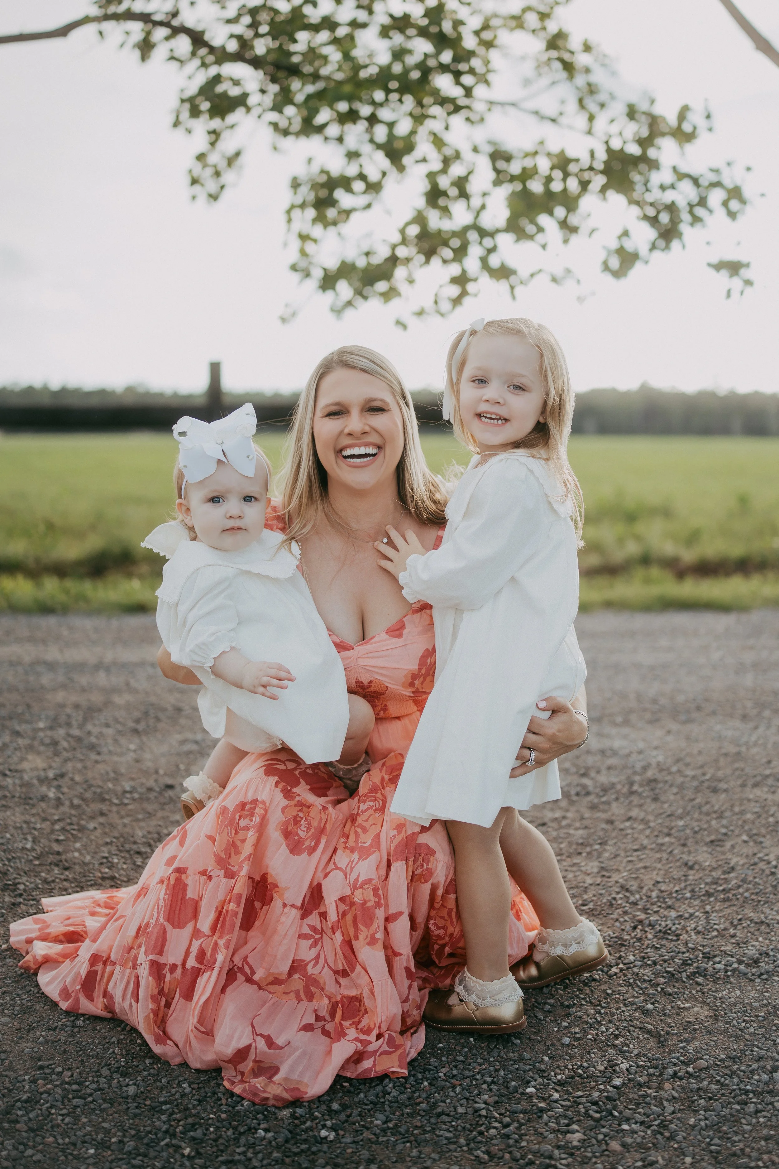 A mother embraces her two young daughters while smiling beneath soft evening light, creating a joyful outdoor family portrait filled with warmth, laughter, and the sweet bond between sisters.