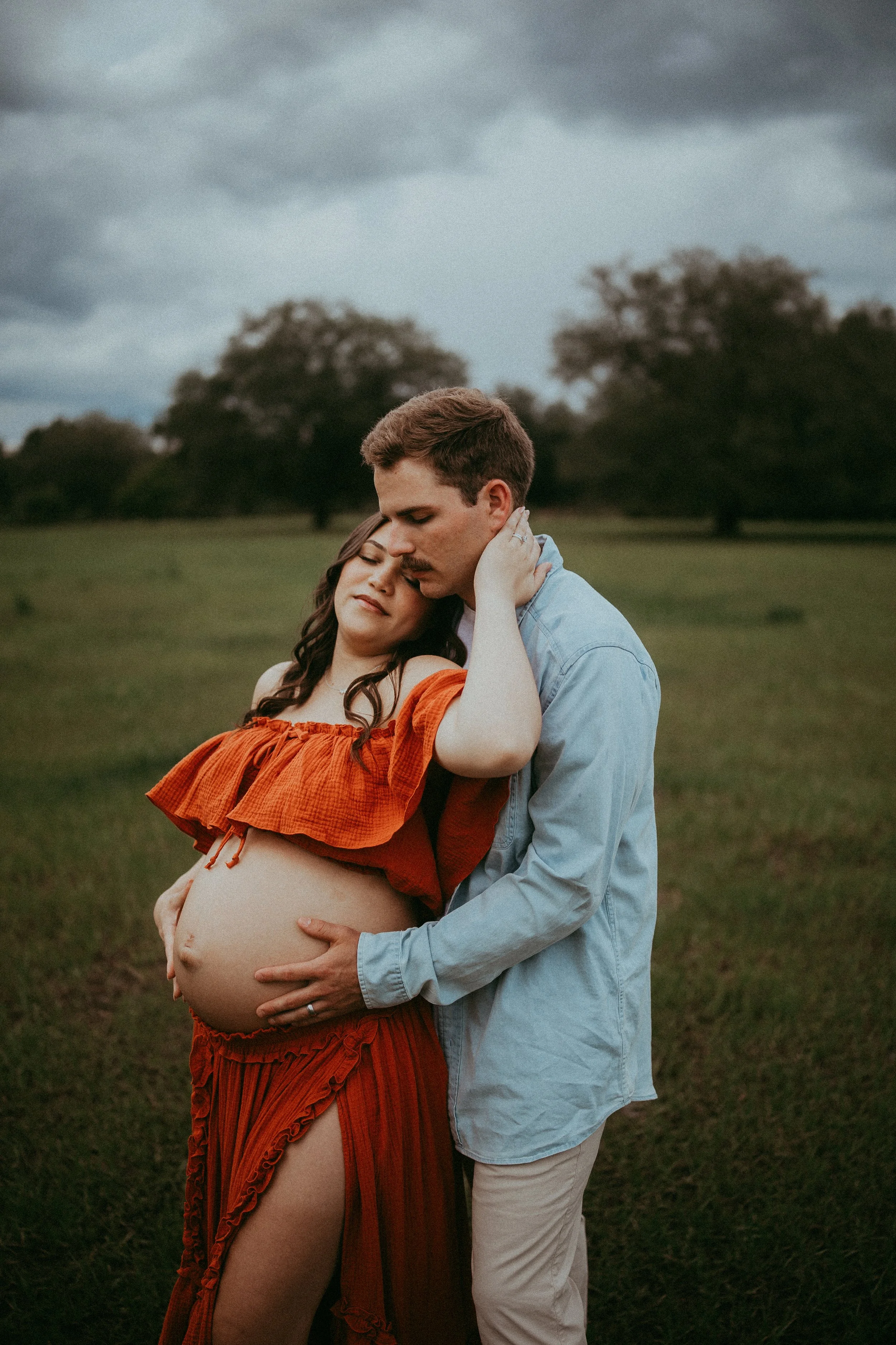 Expectant couple embraces in a grassy field during a romantic maternity session in Hilliard, Florida beneath dramatic storm clouds.