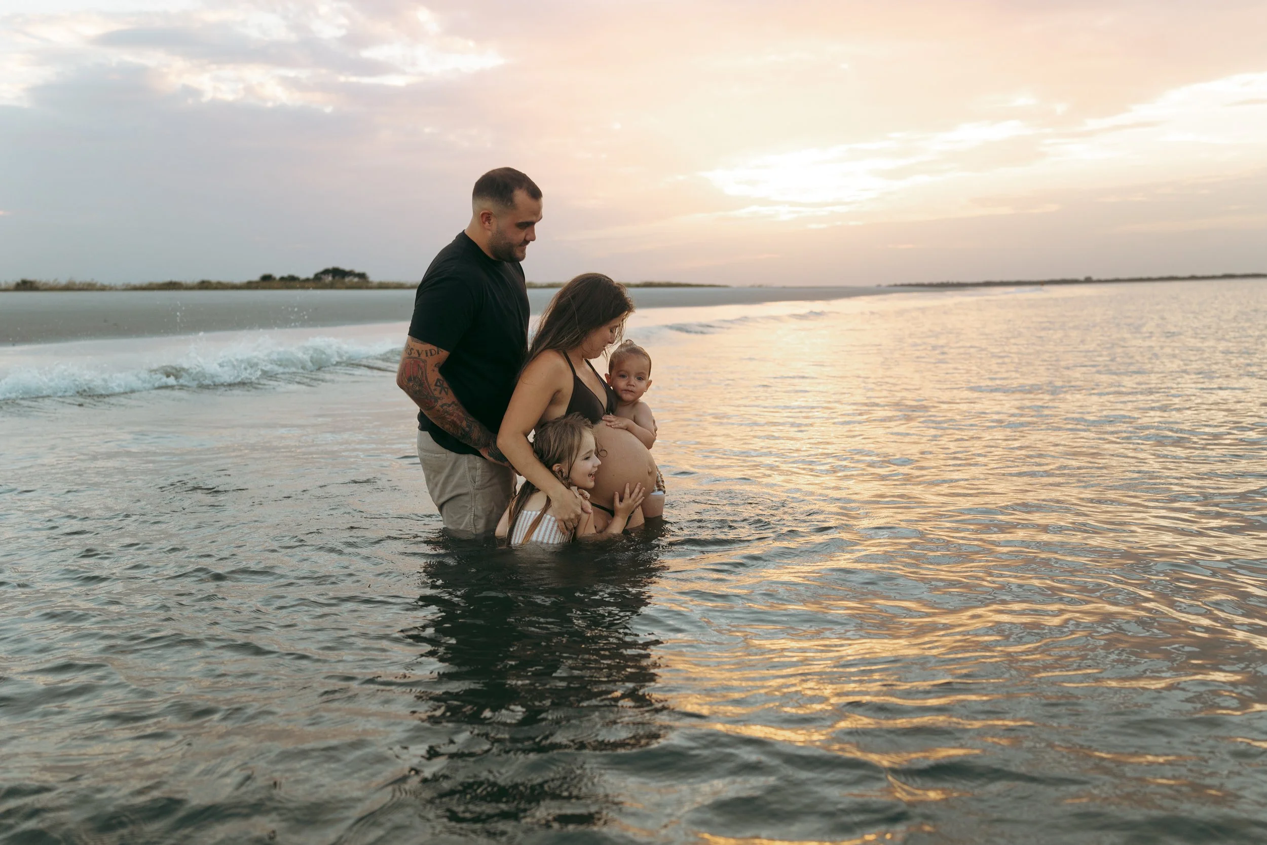 Pregnant mother stands in shallow ocean water with her husband and two young children on Amelia Island Beach at sunset as the family gathers around her baby bump, celebrating the anticipation of welcoming their newest member.