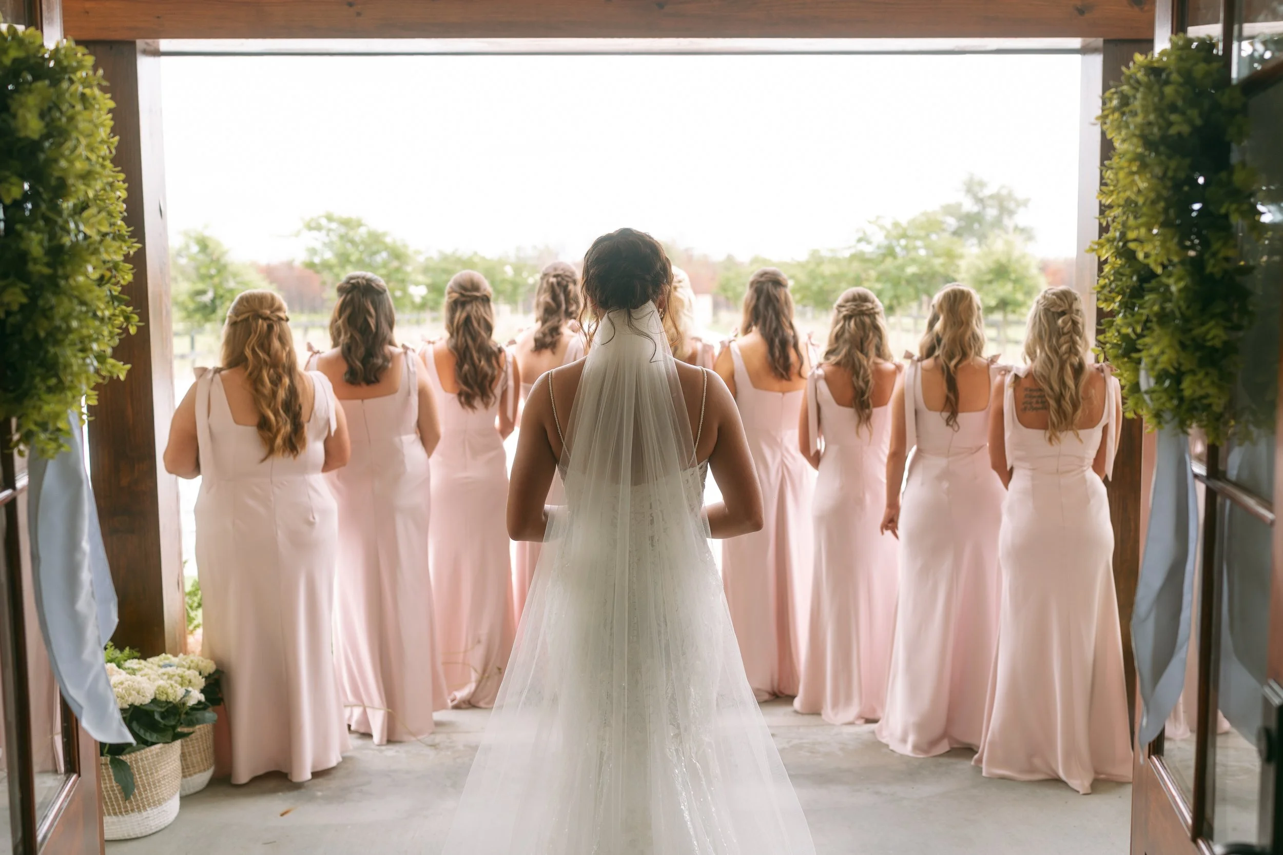 Emma stands behind her bridesmaids in soft blush dresses as they look toward the open field at Red River Estate in Waynesville, Georgia.