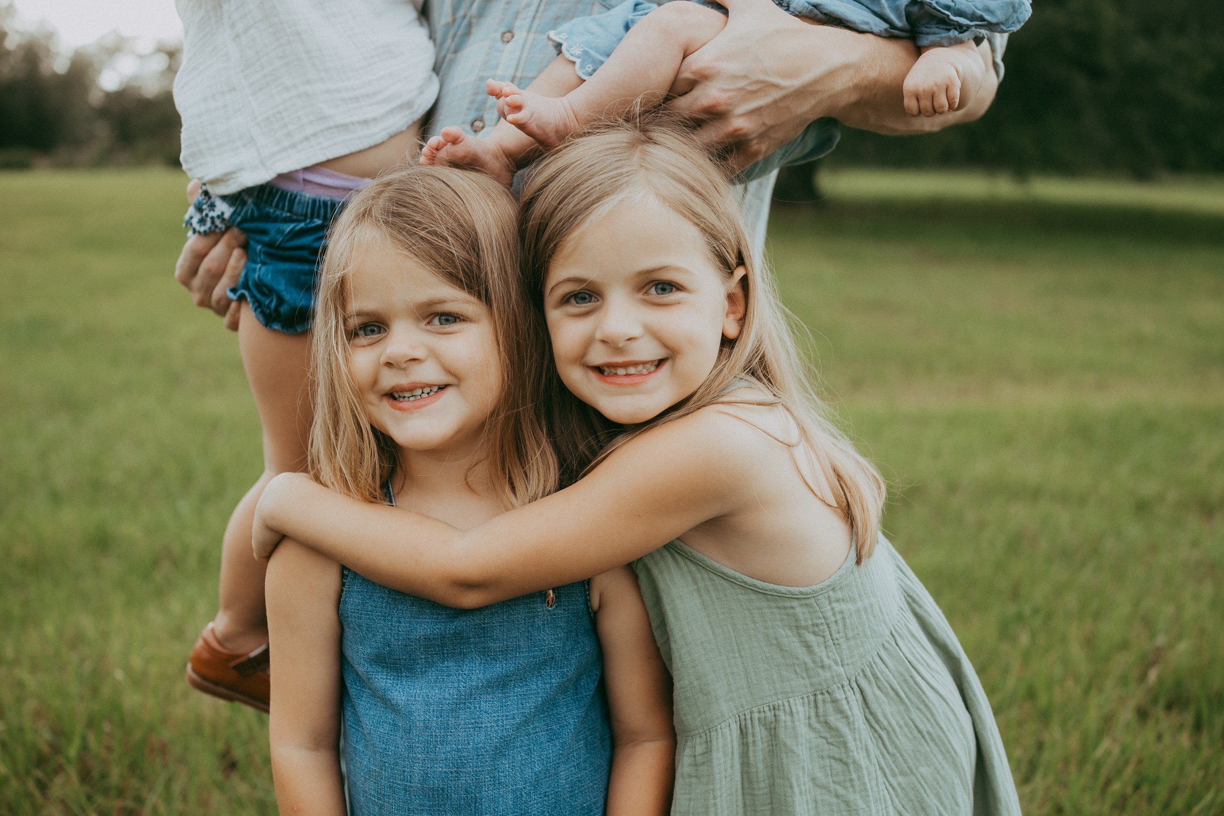Two young sisters share a joyful hug and smile at the camera while standing together in a grassy field, capturing the sweet bond between siblings during a relaxed family photography session.