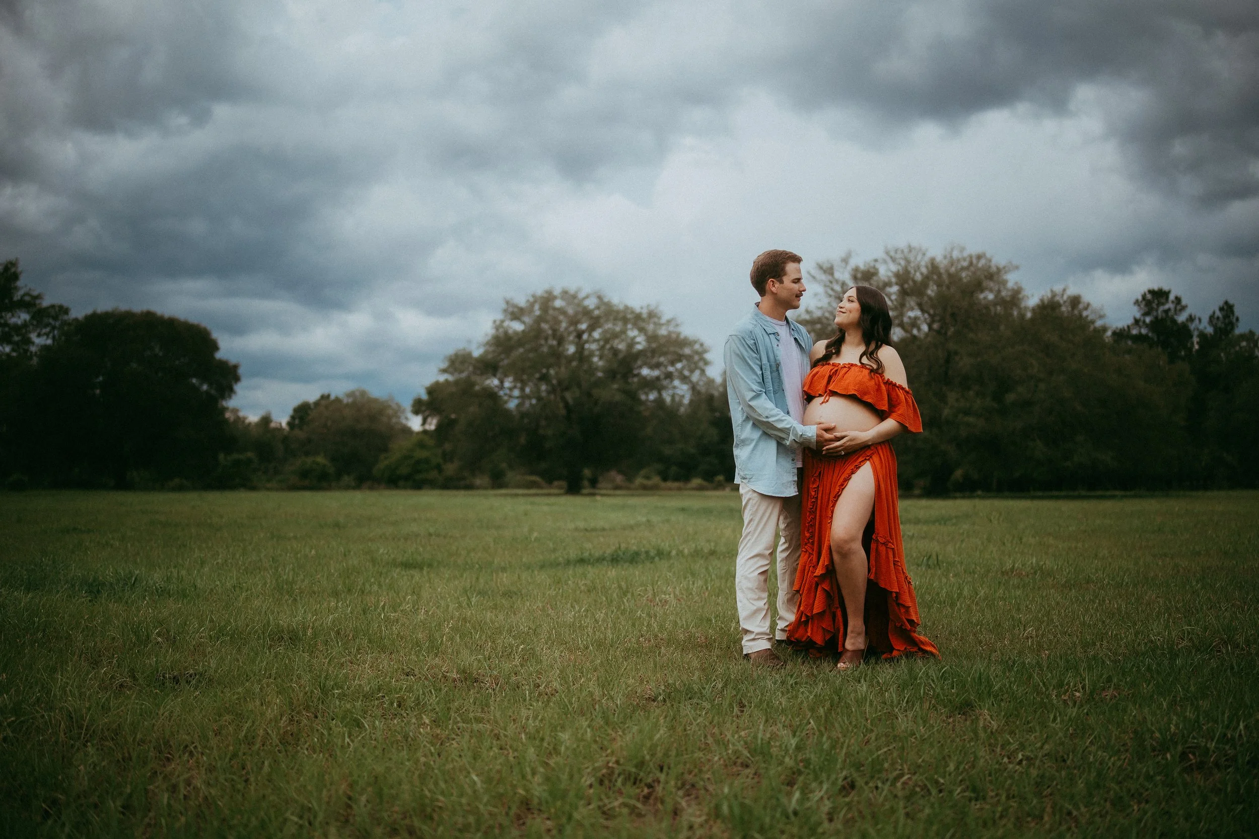 Expectant parents stand together in a grassy field in Hilliard, Florida beneath dramatic storm clouds, gently holding their baby bump during a romantic outdoor maternity session.