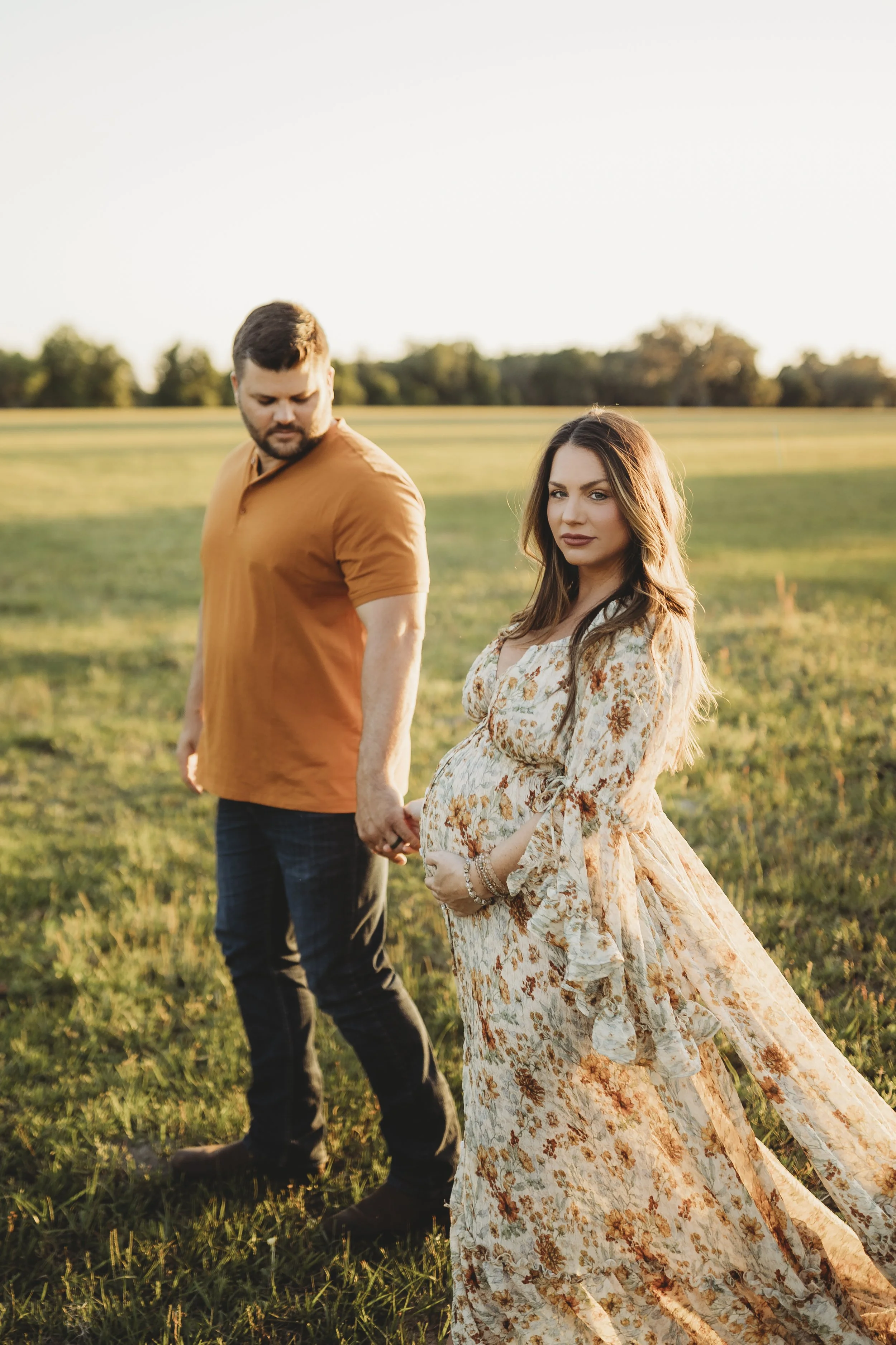 Outdoor maternity portrait of an expecting couple holding hands in an open field at sunset as the mother gently holds her baby bump.
