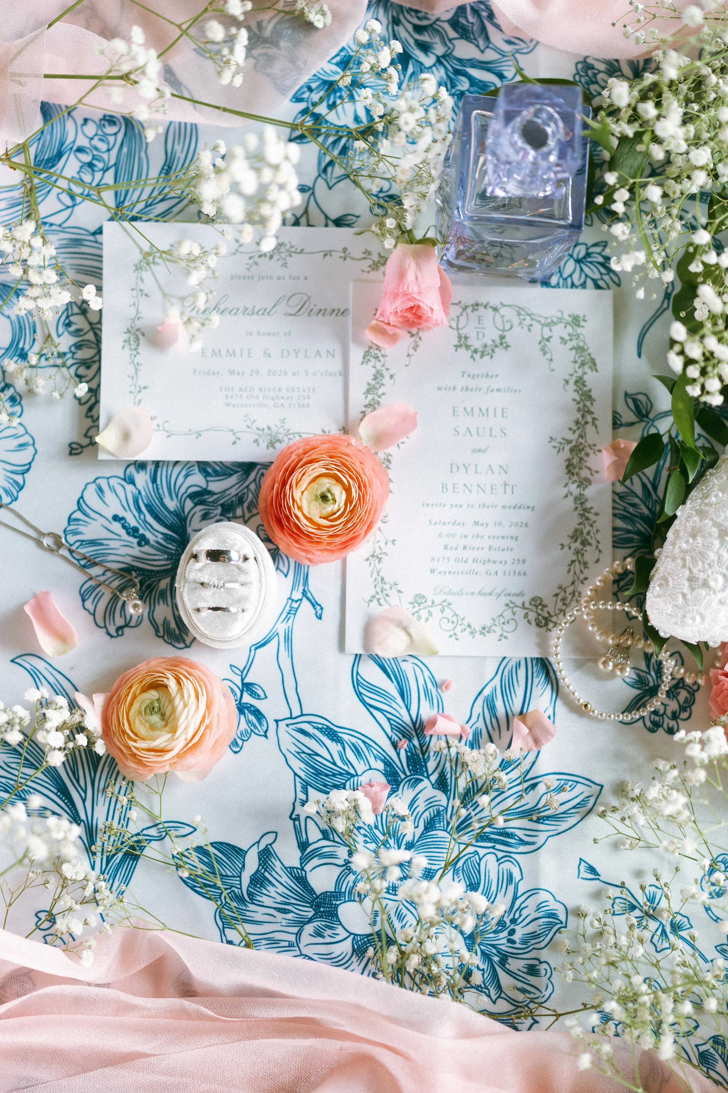Flat lay of Emmie and Dylan’s wedding invitations, rings, perfume, pearls, and flowers for their Red River Estate wedding in Waynesville, Georgia.
