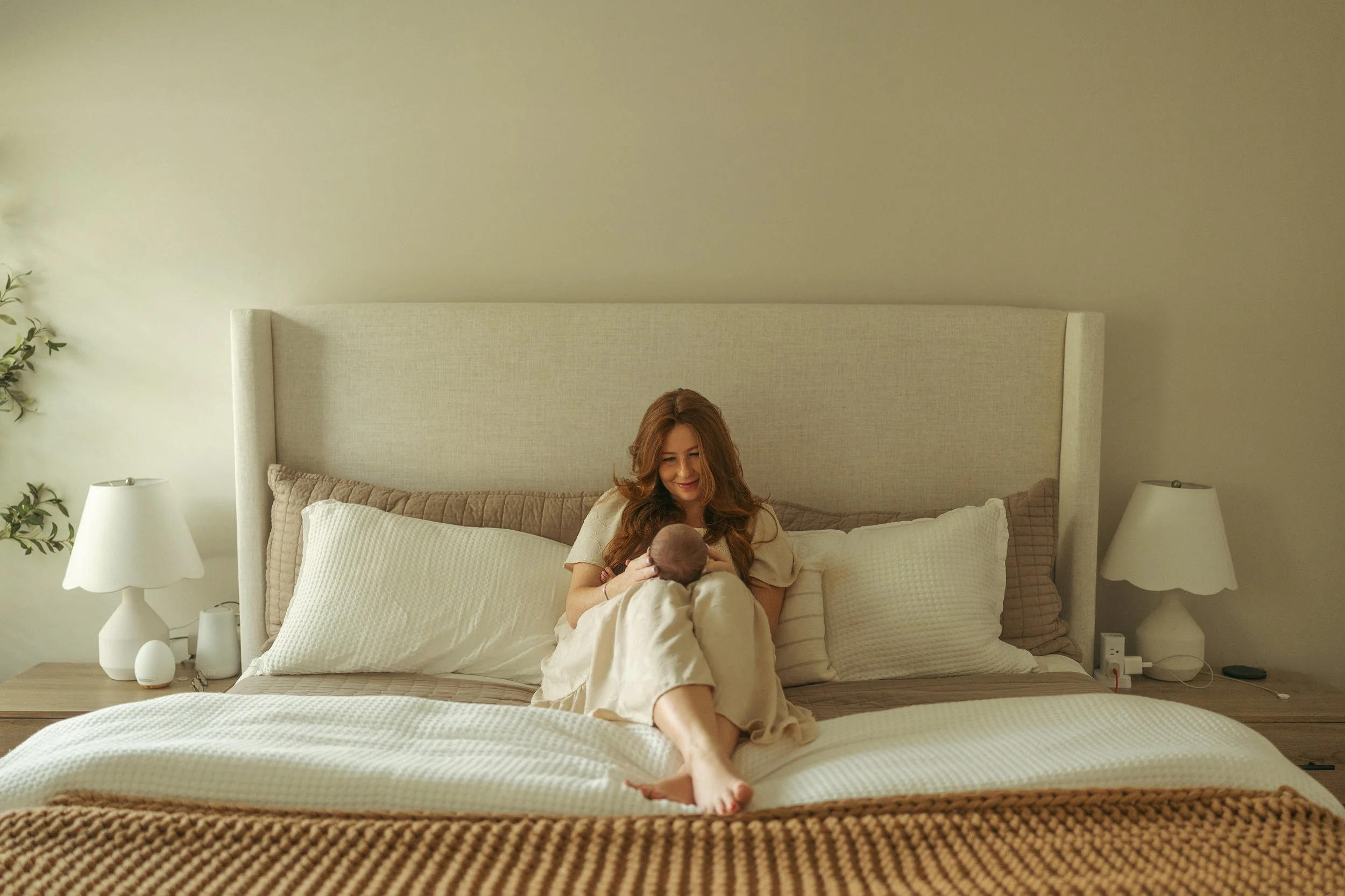 Mother sits on a softly made bed in a light-filled bedroom, smiling down at her newborn cradled in her arms during an intimate in-home lifestyle newborn photography session.
