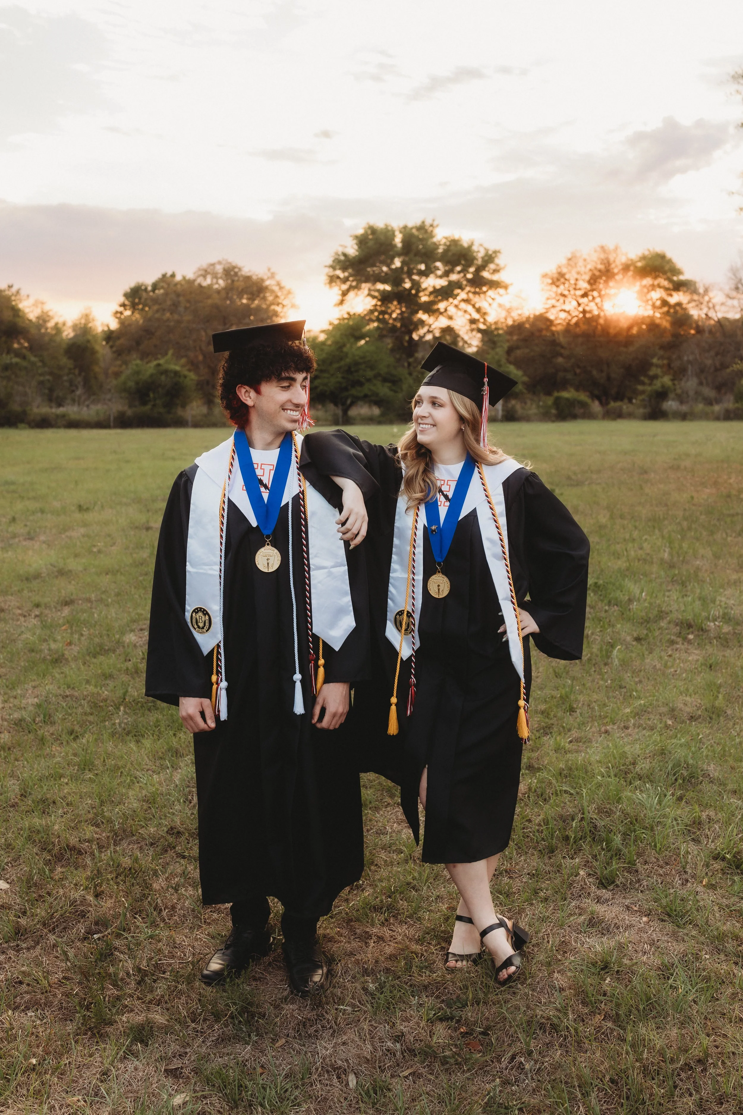 Twin high school seniors smiling together in caps and gowns during an outdoor graduation photography session at sunset in Northeast Florida.