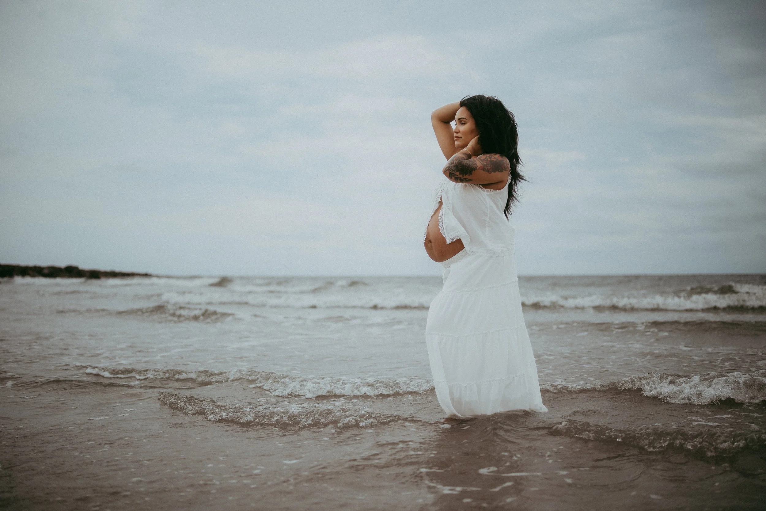 Expectant mother standing in the ocean at sunset wearing a flowing white dress during a beach maternity session, celebrating the beauty of pregnancy and motherhood.