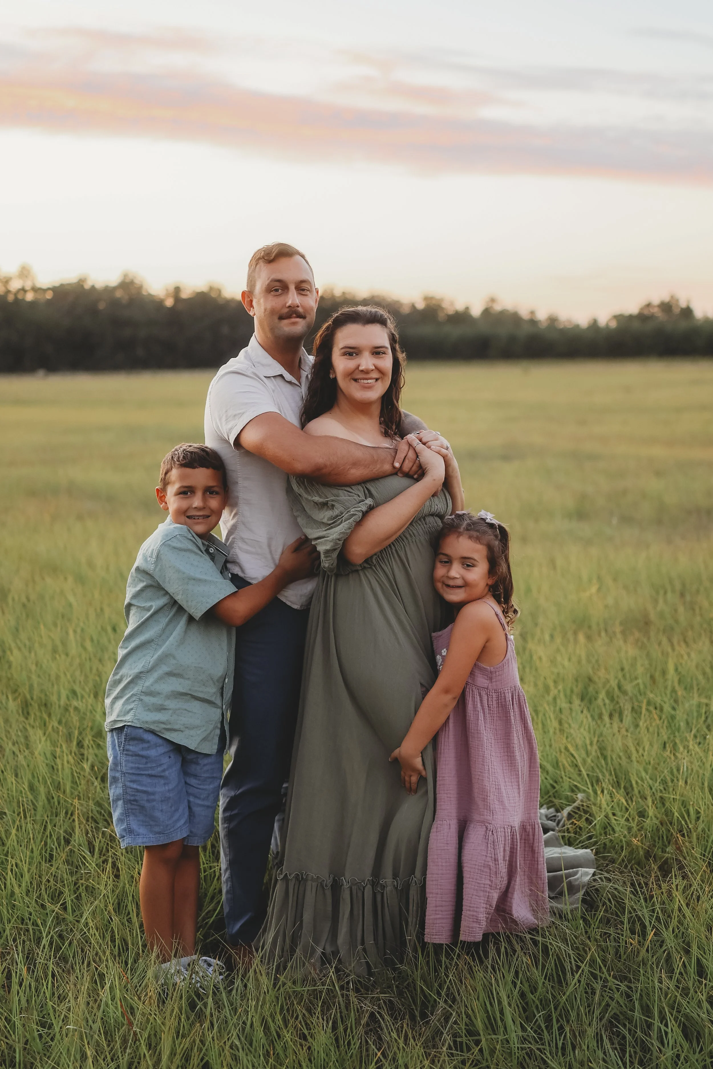 A family of four stands together in a grassy field at sunset, with their son and daughter hugging their parents as they eagerly await the arrival of their newest little one.