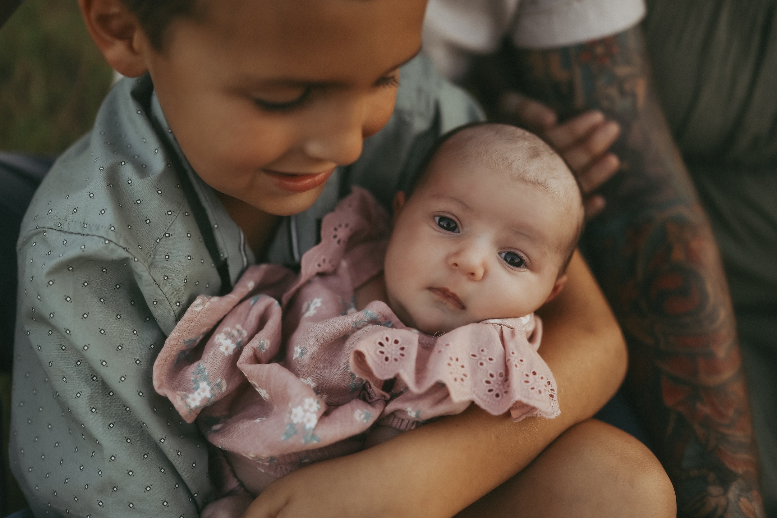 A proud big brother gently holds his newborn sister during a warm outdoor family session at sunset.