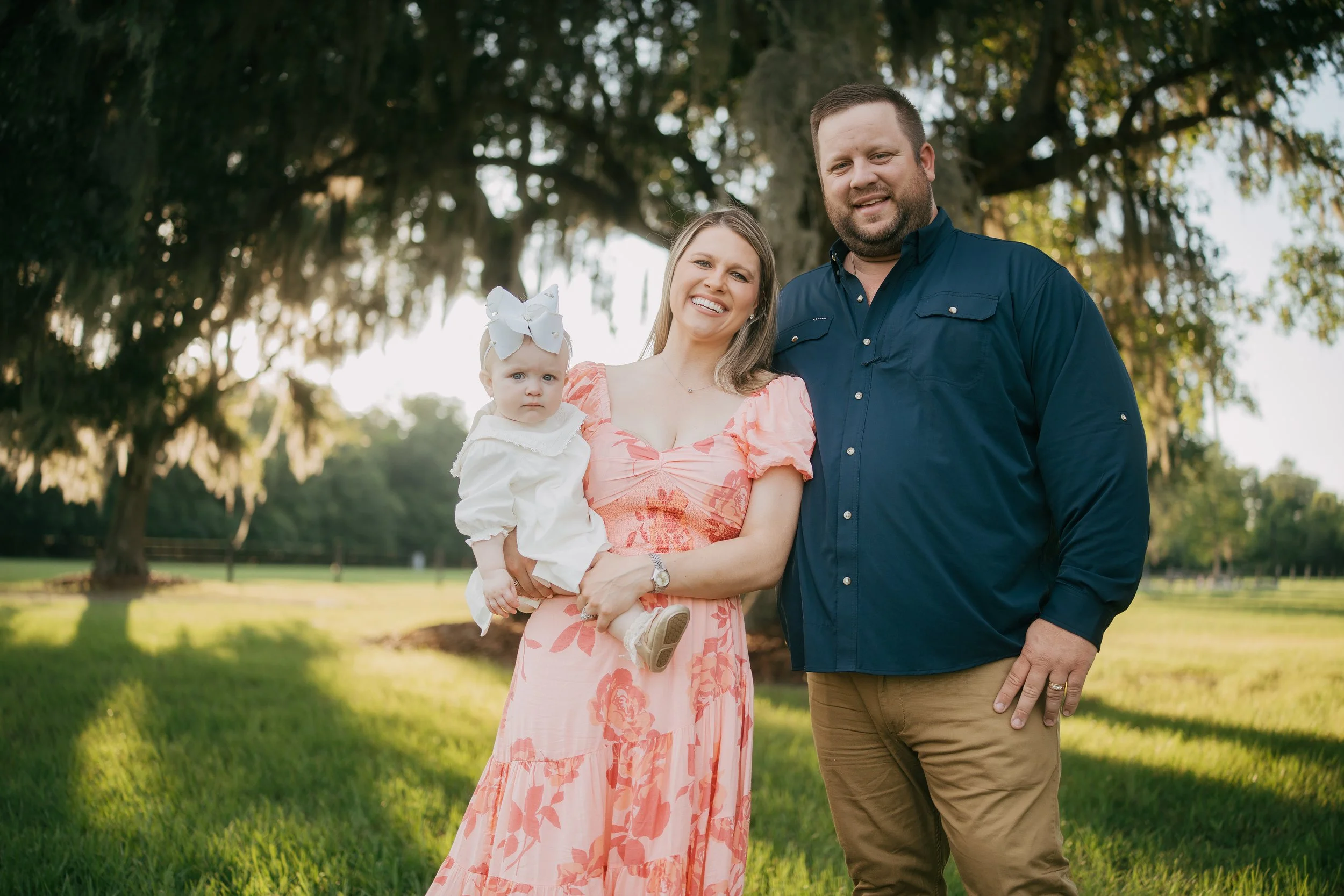 A smiling family of three stands beneath majestic oak trees during golden hour, with parents holding their baby girl for a timeless outdoor family portrait filled with warmth and love.