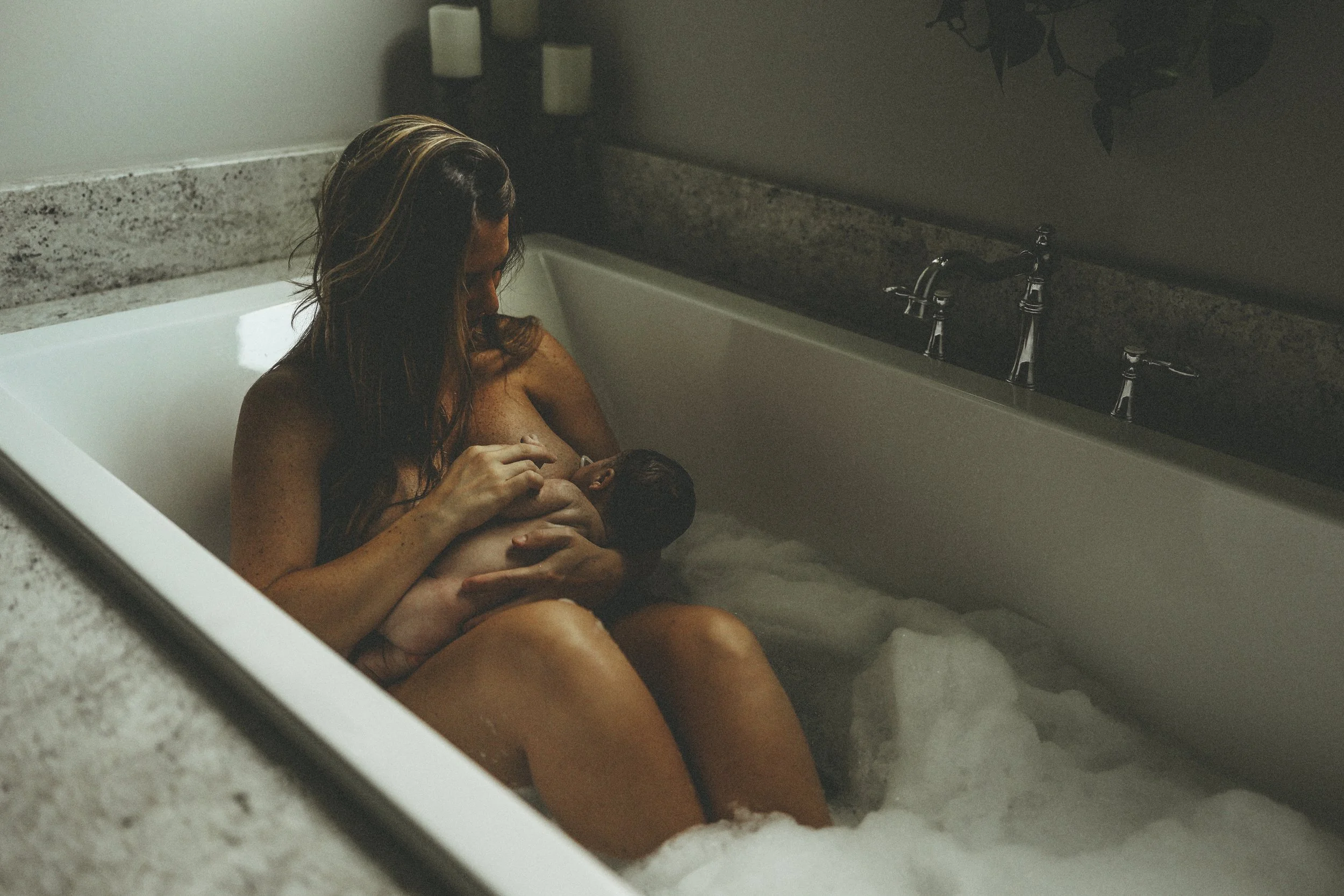 Parent sitting in a bathtub holding and nursing a newborn, surrounded by soft bathwater and gentle light.