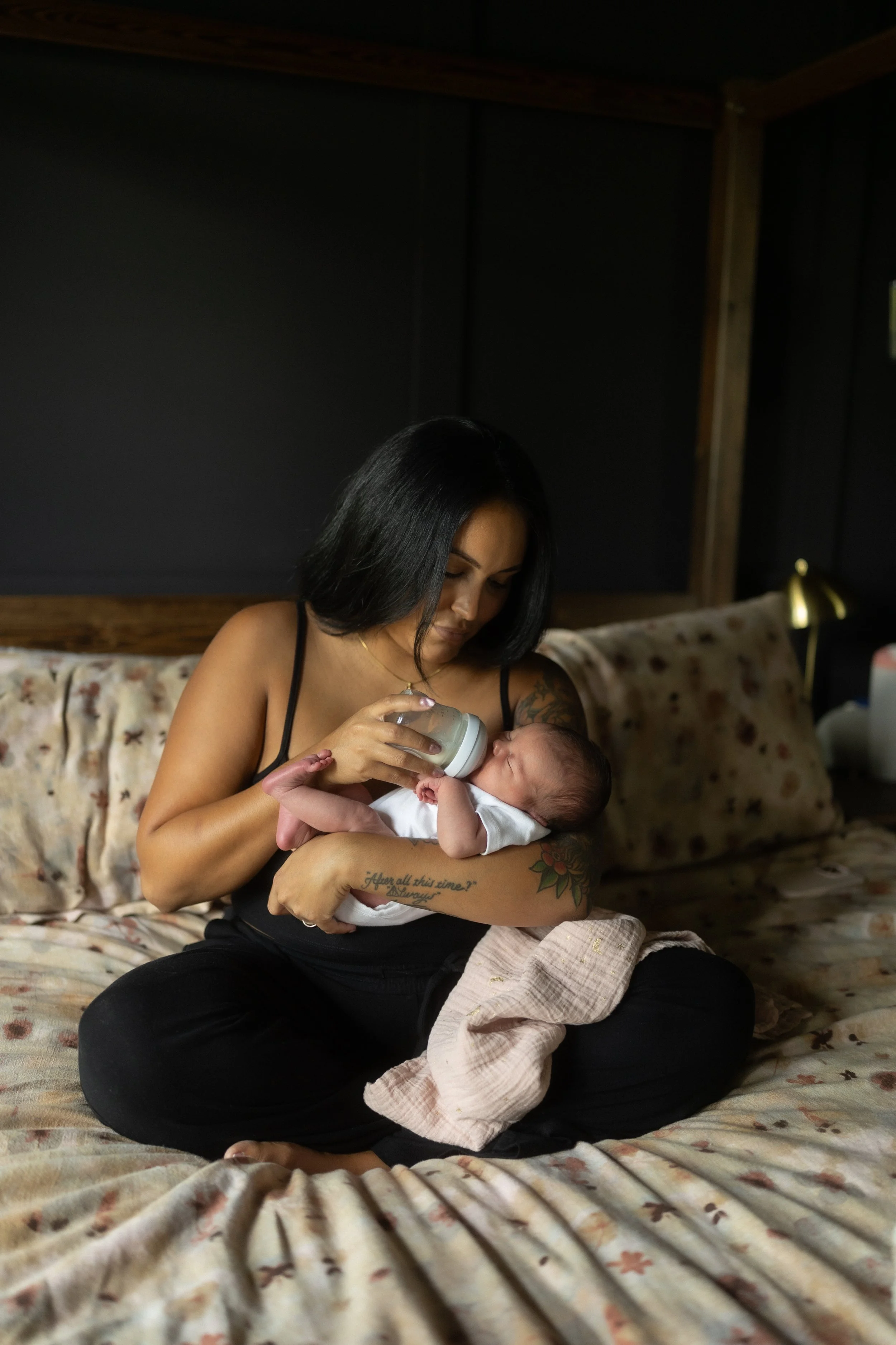 Mother bottle-feeds her newborn while sitting cross-legged on the bed, creating a peaceful everyday moment that reflects the love and care of an in-home lifestyle newborn photography session.
