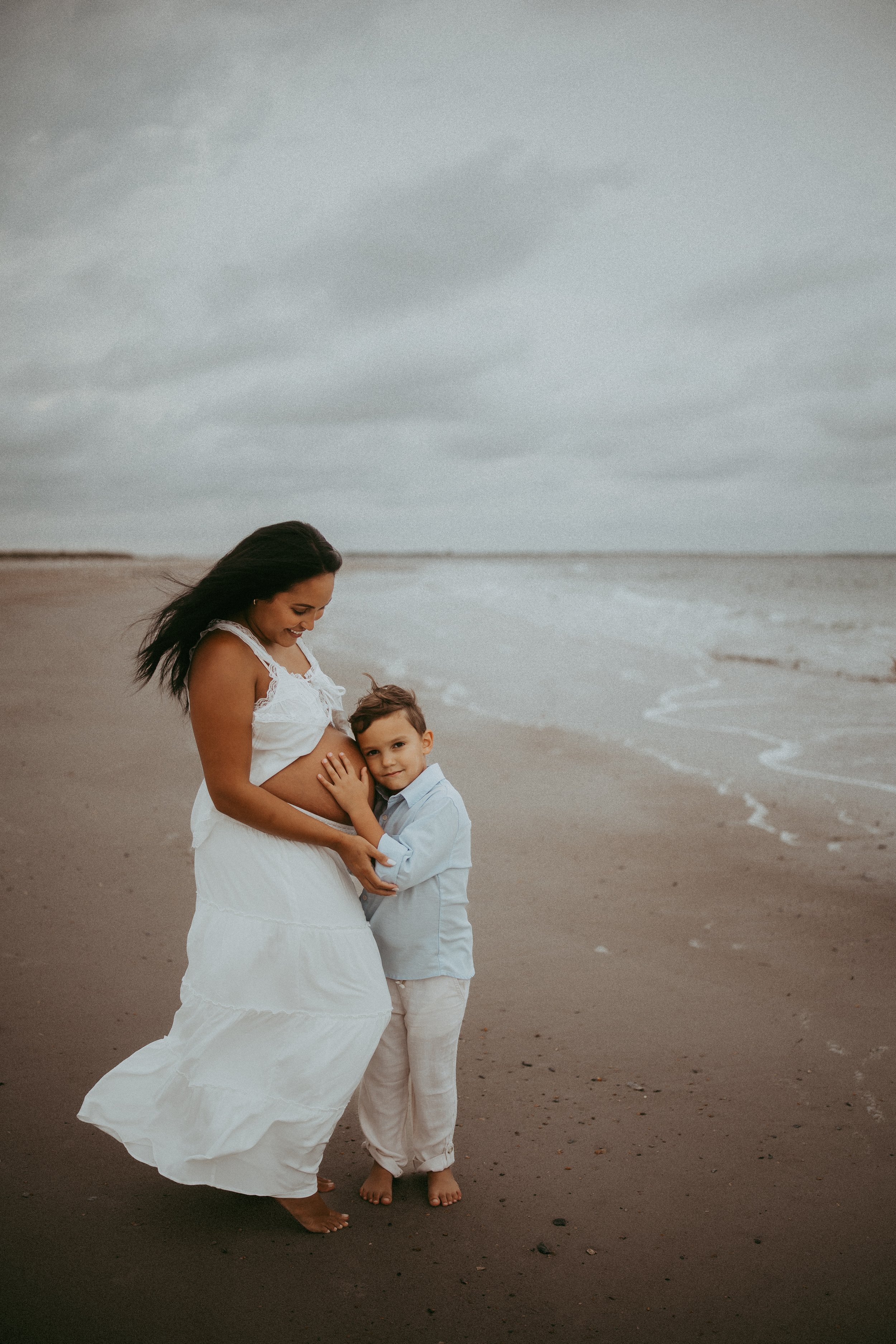 Young boy hugs his mother's pregnant belly on the beach at Fort Clinch State Park in Fernandina Beach, Florida, as they share a sweet moment of anticipation during a relaxed beach maternity photography session.