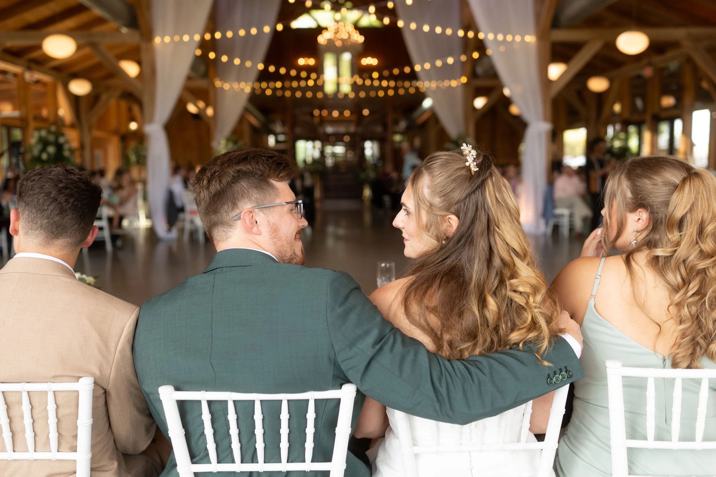 The newlyweds smile at one another while seated together during their wedding reception.