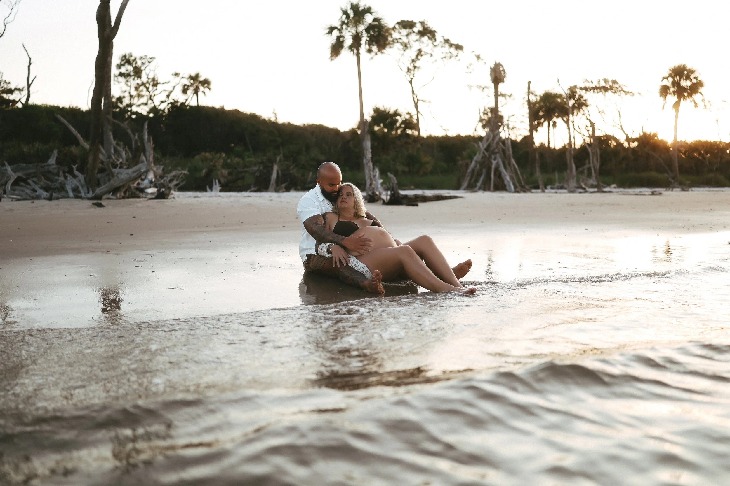 Sunset maternity photo of an expectant couple sitting along the shoreline, embracing peacefully as the tide moves around them.