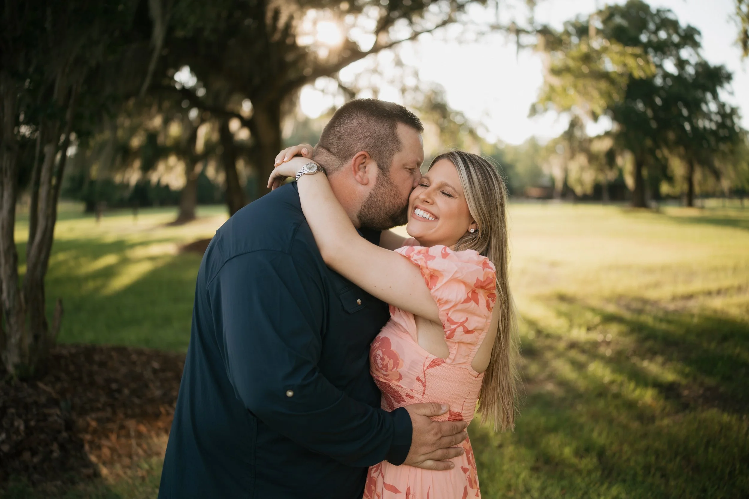 A husband kisses his smiling wife as they embrace beneath sunlit oak trees, creating a heartfelt moment of love and connection during their outdoor family portrait session.