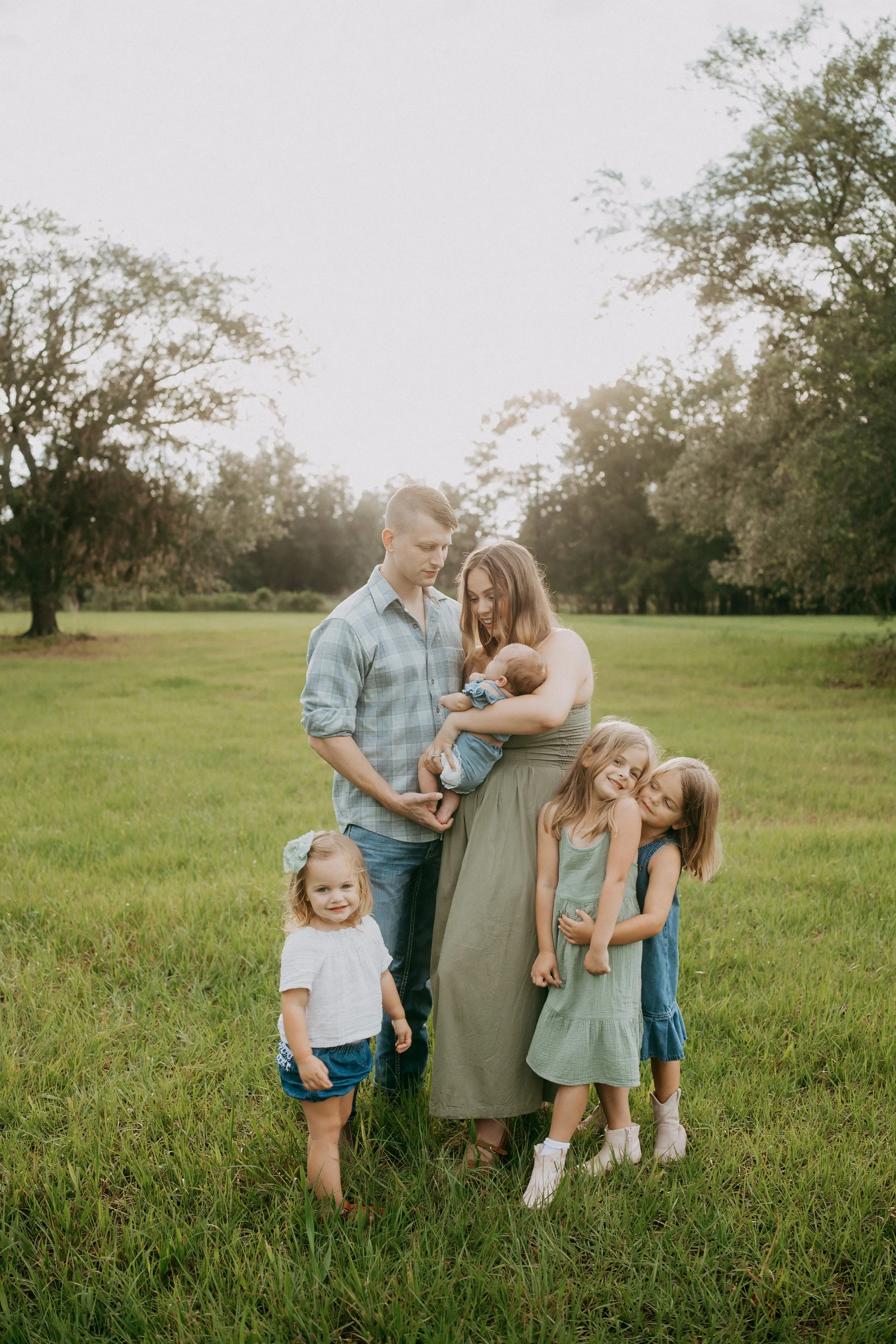 A family of six stands together in a peaceful grassy field as the parents admire their newborn, two sisters share a sweet hug, and their youngest daughter smiles proudly at the camera, capturing the joy of everyday family life.