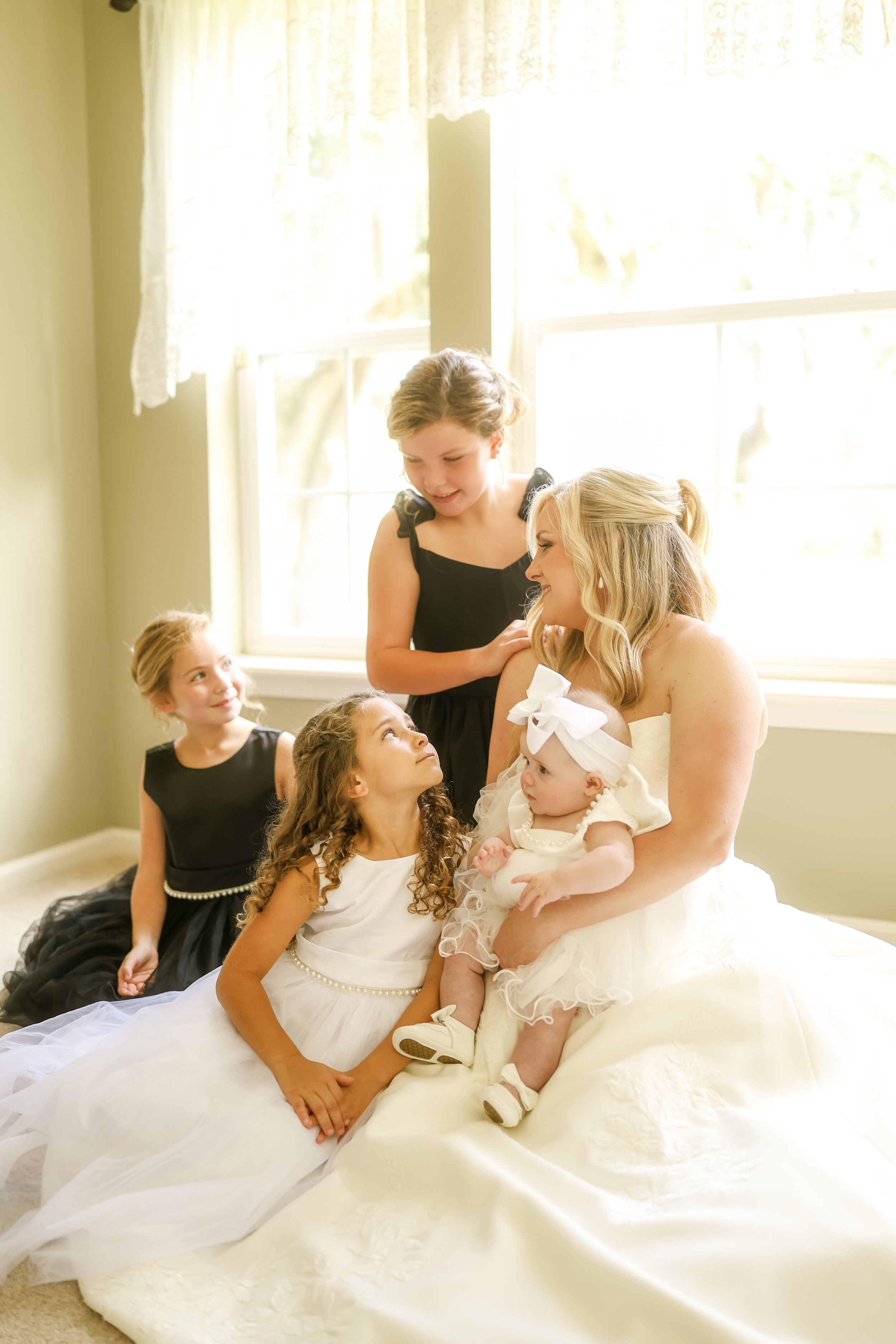 Seated in soft window light, the bride gathers her daughters and stepdaughters around her, a quiet moment where laughter, trust, and a mother’s love come together to tell the story of a family beautifully becoming one.