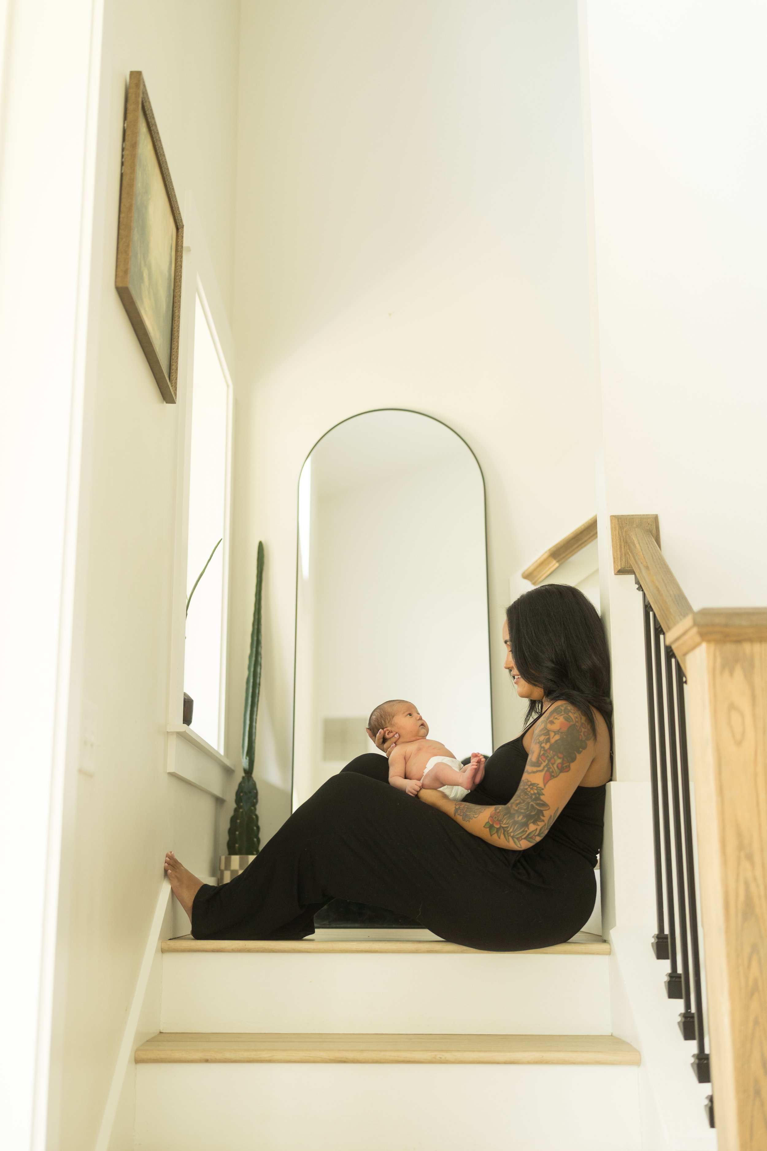Mother sits on a staircase holding her newborn in her arms as they gaze at one another, creating a quiet, light-filled moment during an in-home lifestyle newborn photography session.