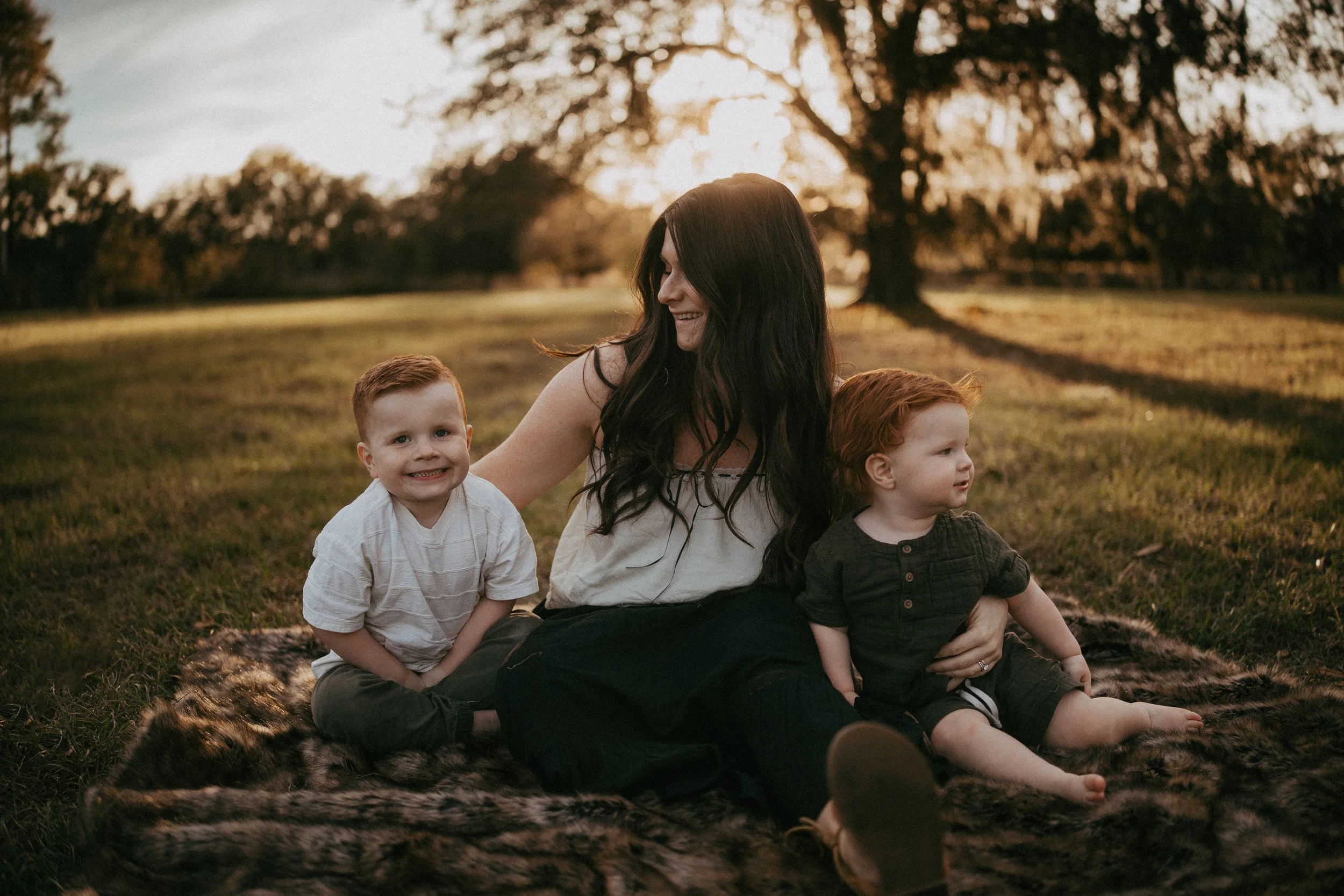 A mother smiles as her young sons sit beside her on a blanket during a relaxed family portrait.