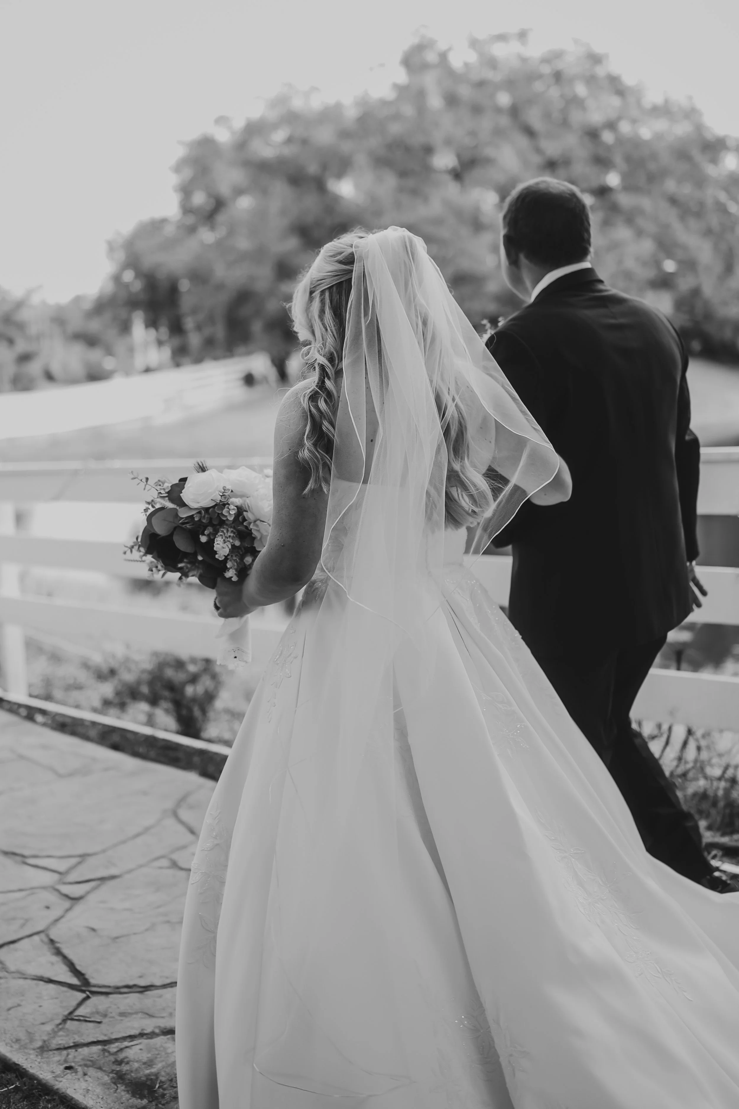 From behind, the bride walks beside her father, her veil and gown flowing softly as they share a quiet, meaningful moment on the way to the ceremony.