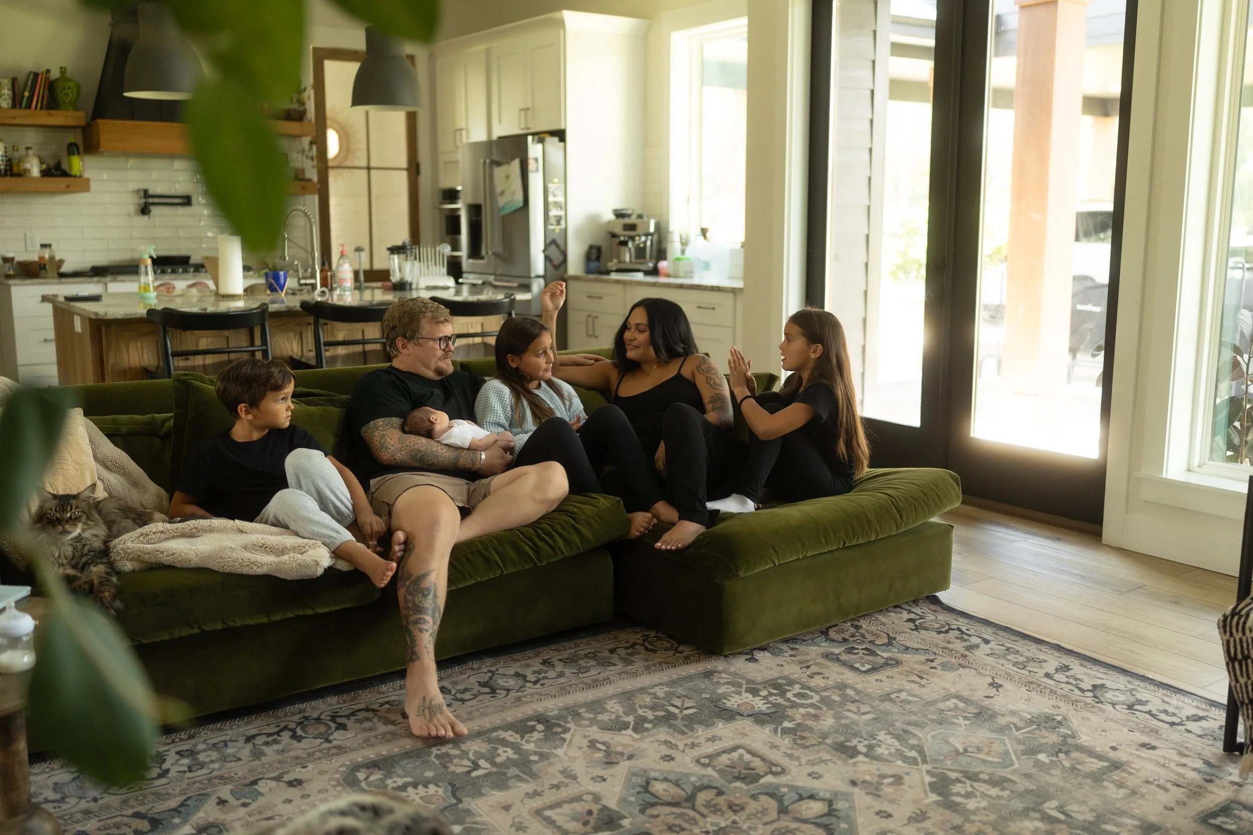 Family of six relaxes together on a cozy living room sofa with their newborn, sharing conversation, laughter, and everyday moments during an in-home lifestyle newborn photography session.