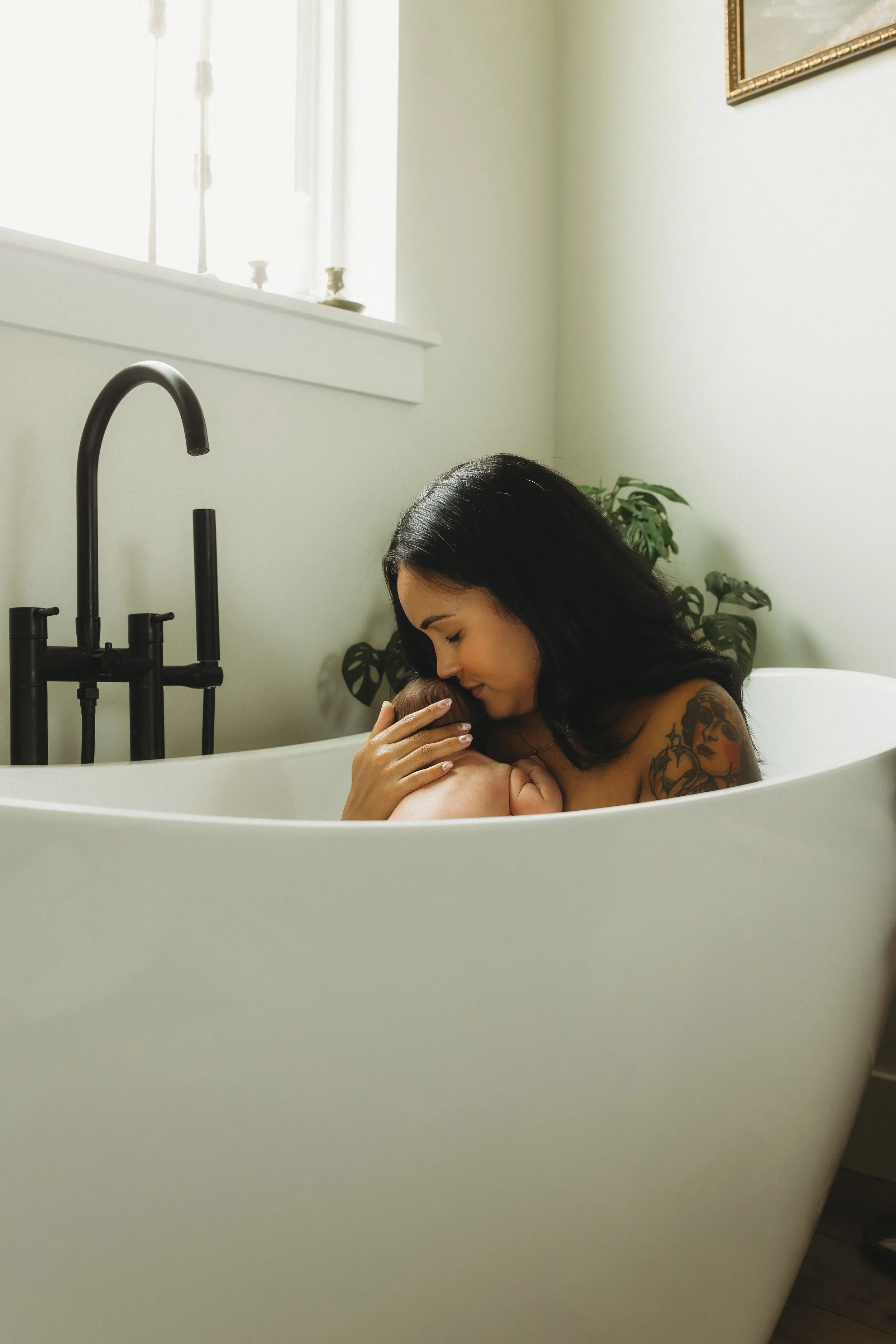 Mother cuddles her sleeping newborn against her shoulder while sitting in a bathtub, capturing a peaceful skin-to-skin inspired moment during an intimate in-home lifestyle newborn photography session.