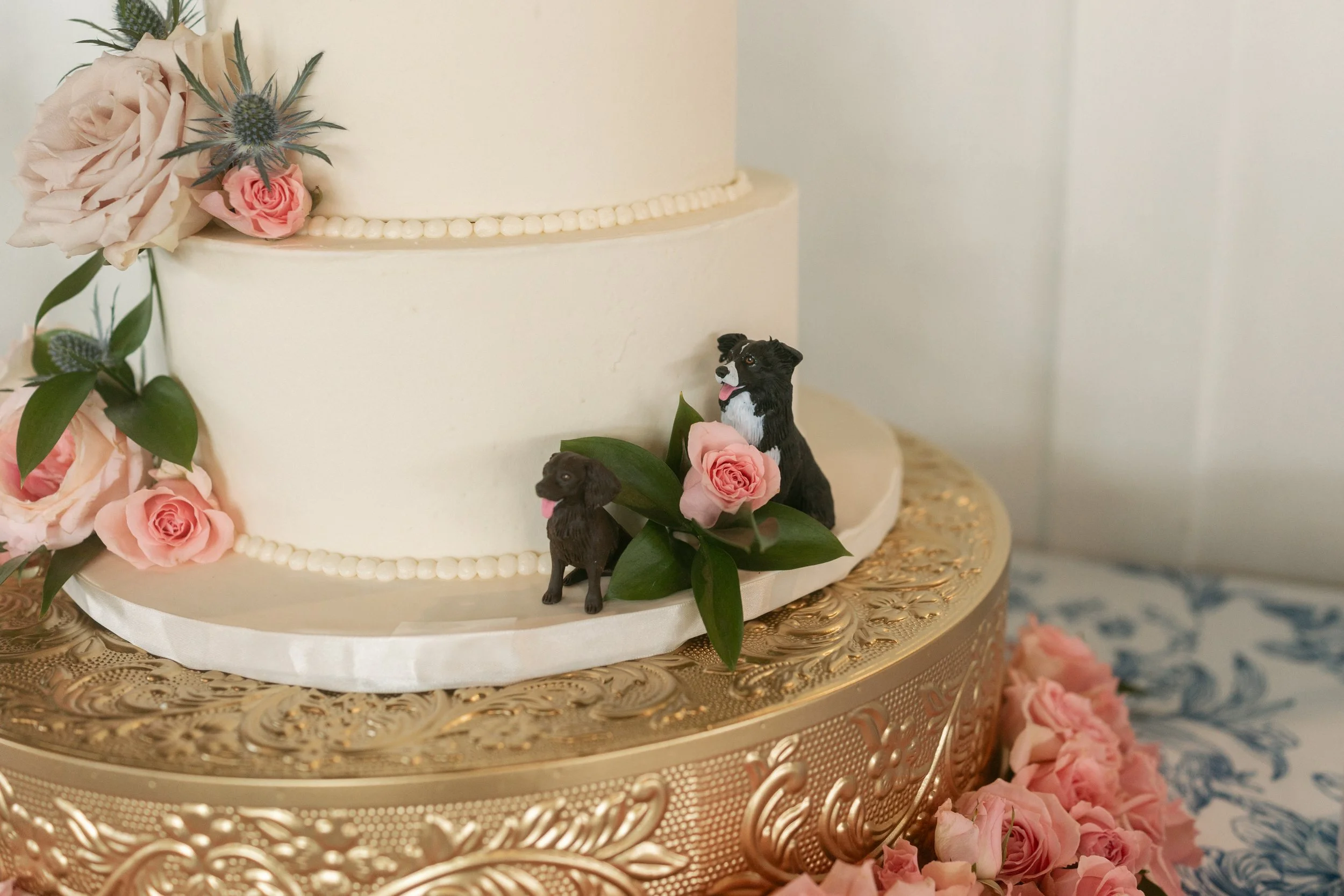 Close-up of Emmie and Dylan’s white wedding cake decorated with blush roses, greenery, and small figurines of their dogs.
