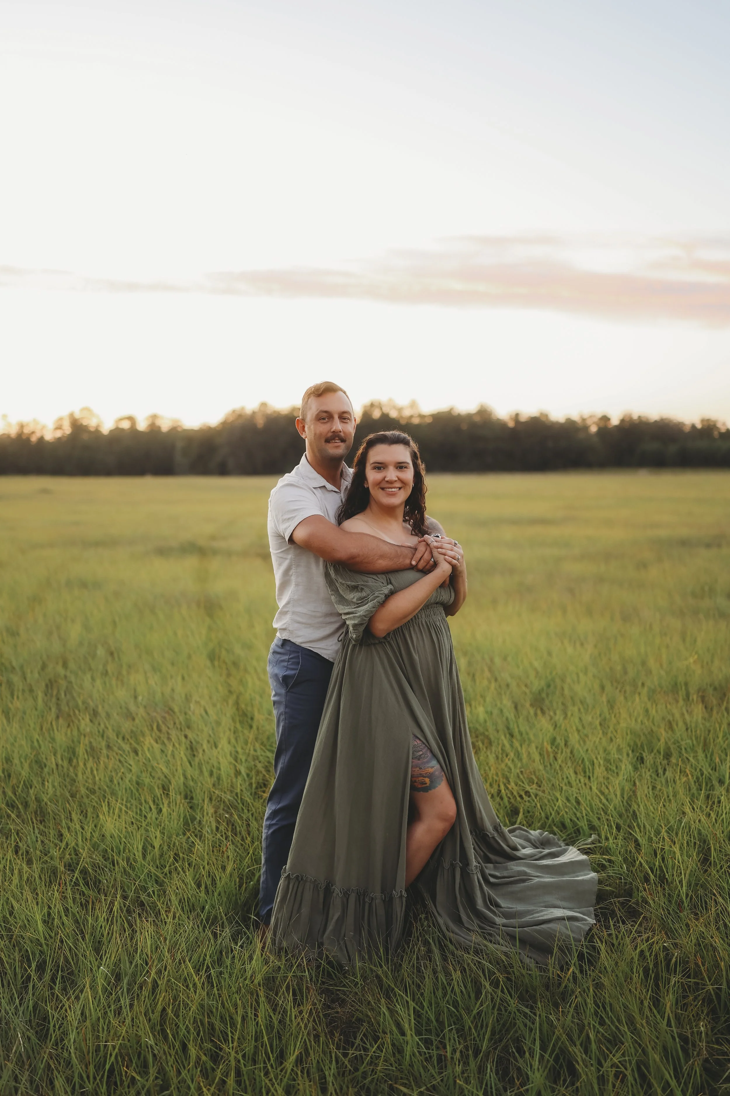 A couple embraces in an open field at sunset, celebrating the anticipation of their growing family during a warm and peaceful maternity session.