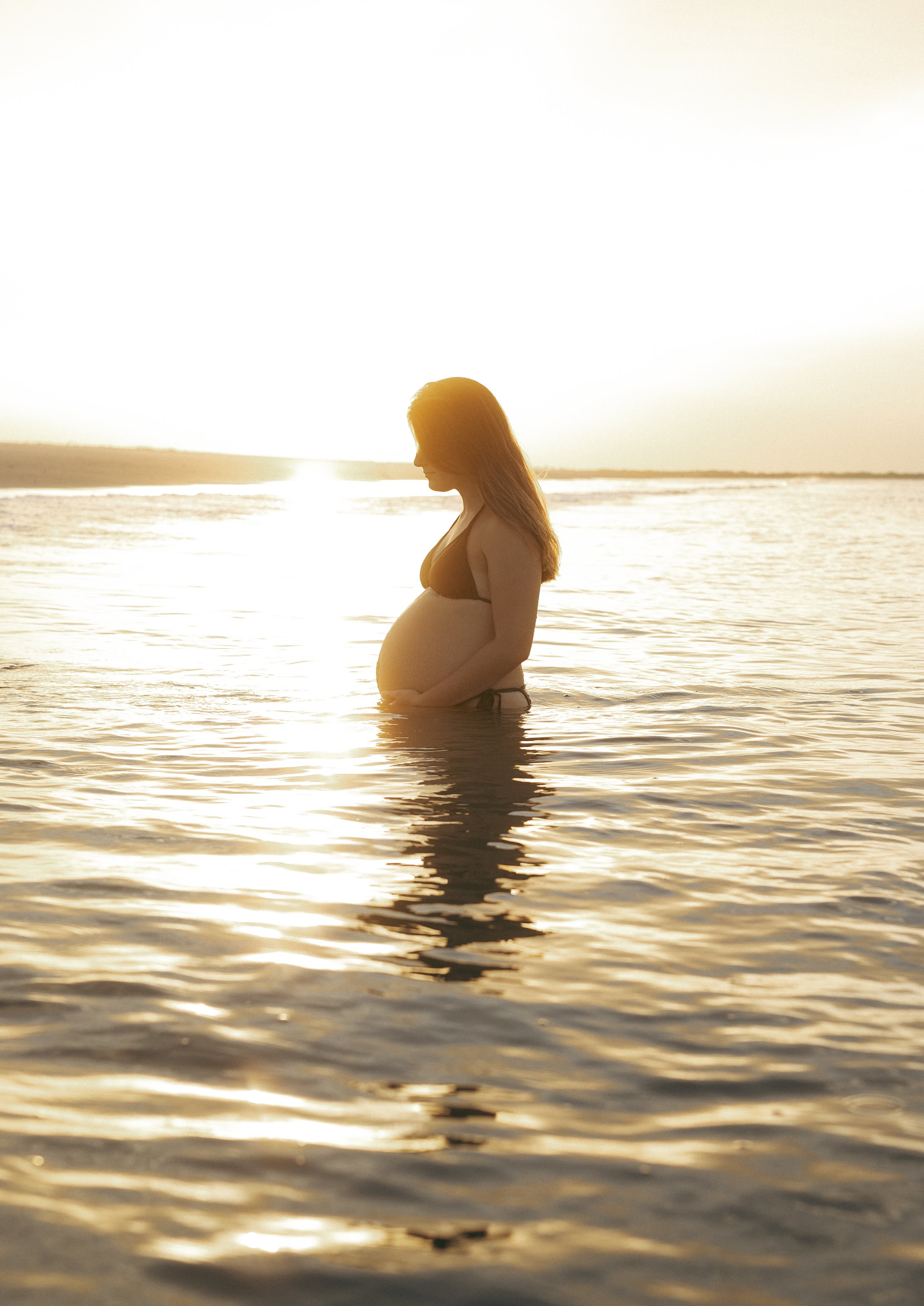 Pregnant mother stands peacefully in calm ocean water at sunset on Amelia Island, silhouetted by golden light while gently holding her baby bump.