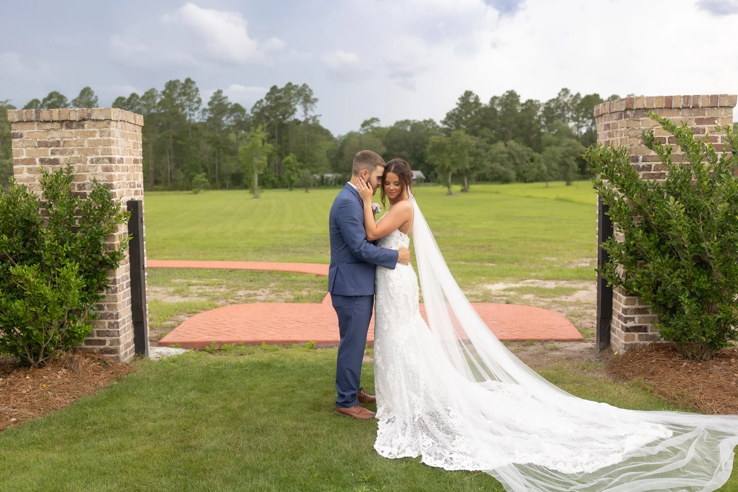 Emma and Dylan embrace between brick pillars as her long veil trails across the lawn beneath a dramatic cloudy sky at Red River Estate.