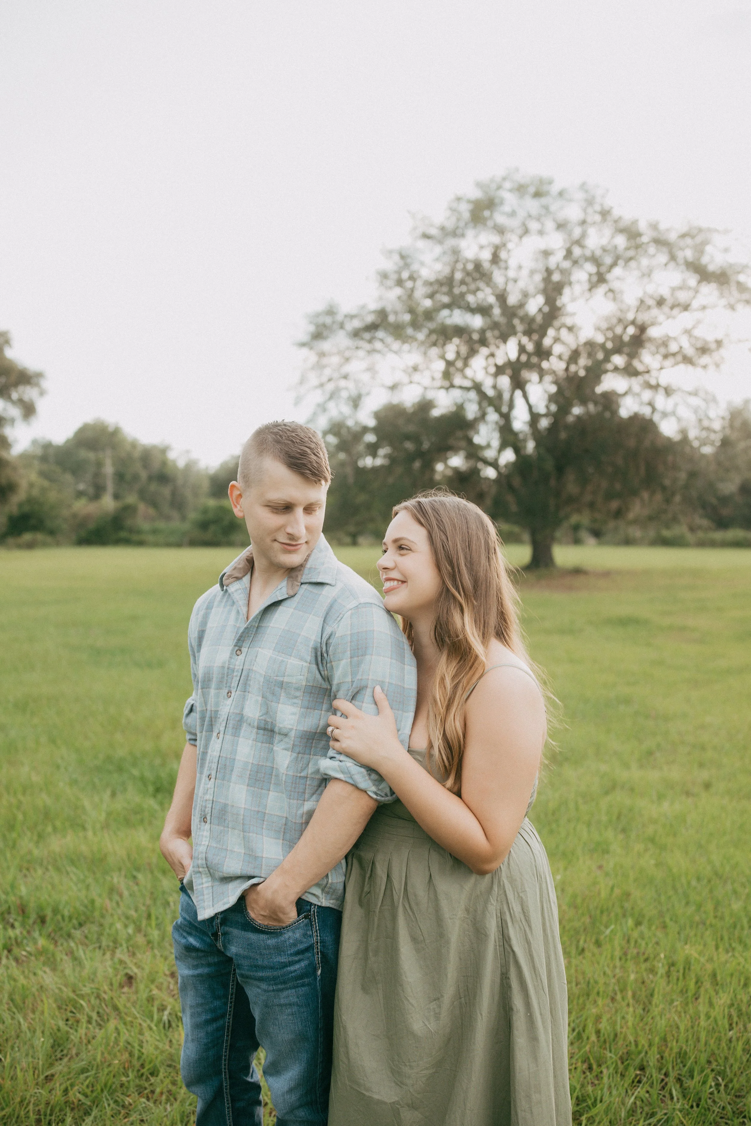 A mother smiles up at her husband as they stand together in a sunlit grassy field, sharing a quiet moment of connection during their family photography session while their children play nearby.
