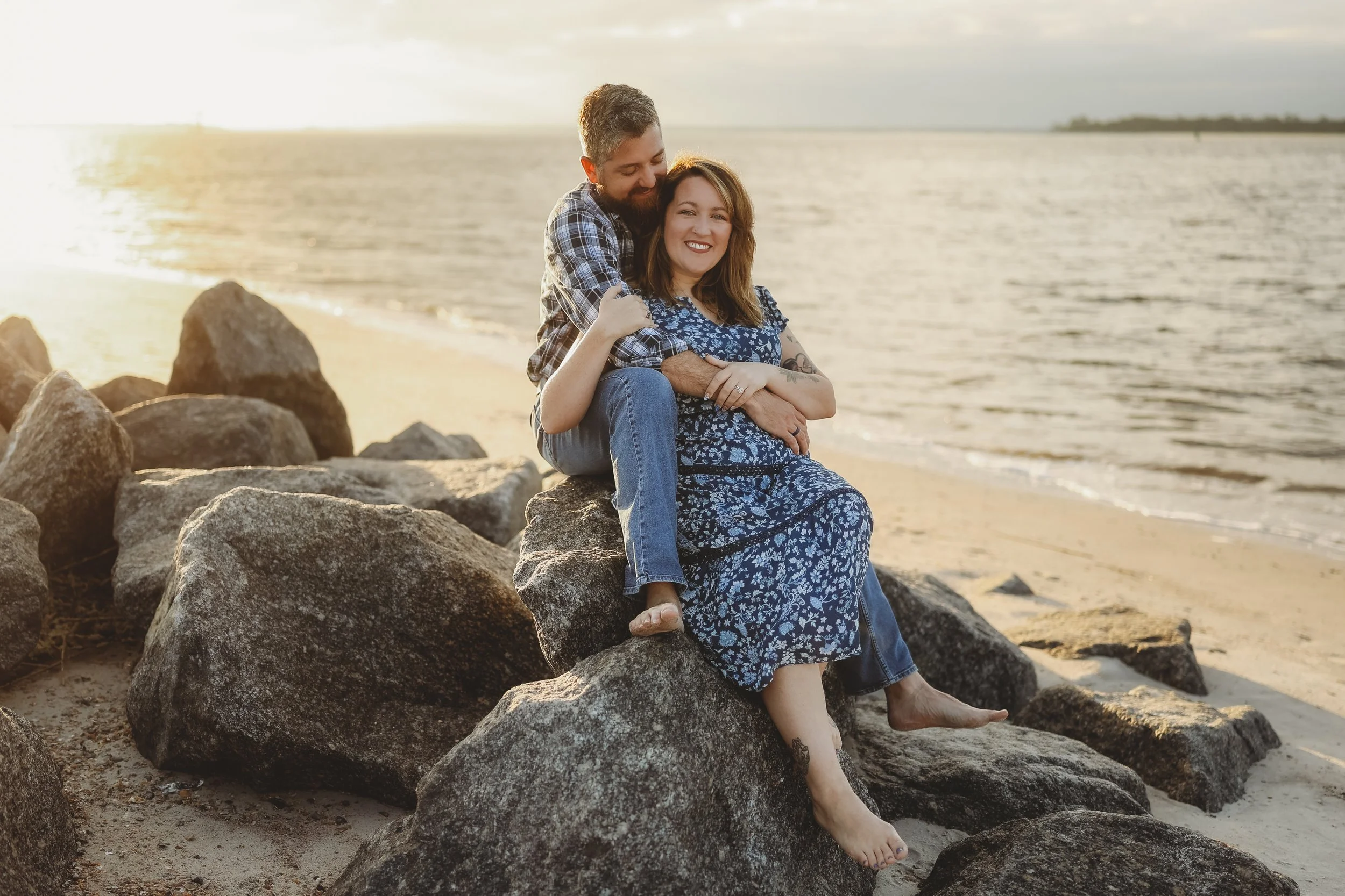 A husband wraps his arms around his wife as they relax on seaside rocks during a golden-hour beach portrait.