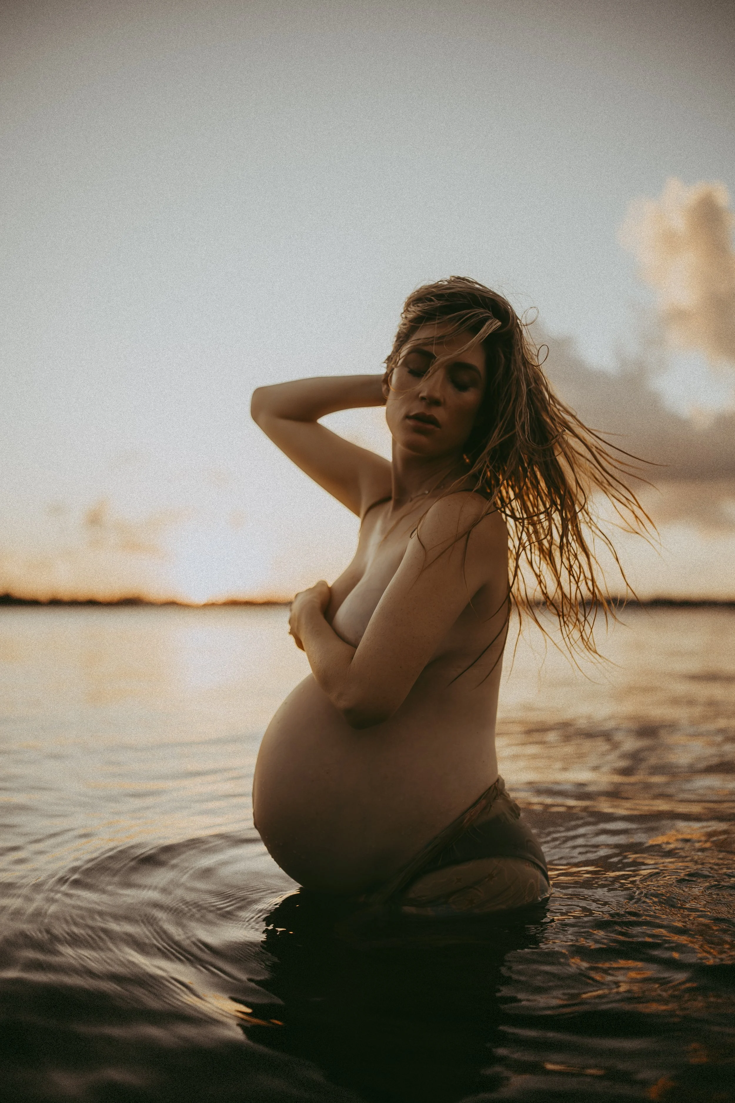 Fine art maternity image of an expectant mother in shallow water at golden hour, capturing vulnerability, strength, and the beauty of motherhood.