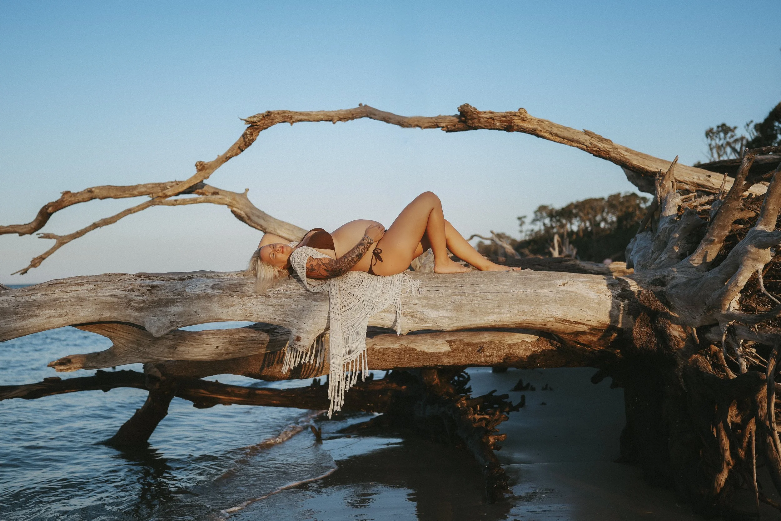 Artistic maternity photo of an expectant mother reclining on driftwood at the beach during golden hour, resting peacefully as the ocean surrounds her.