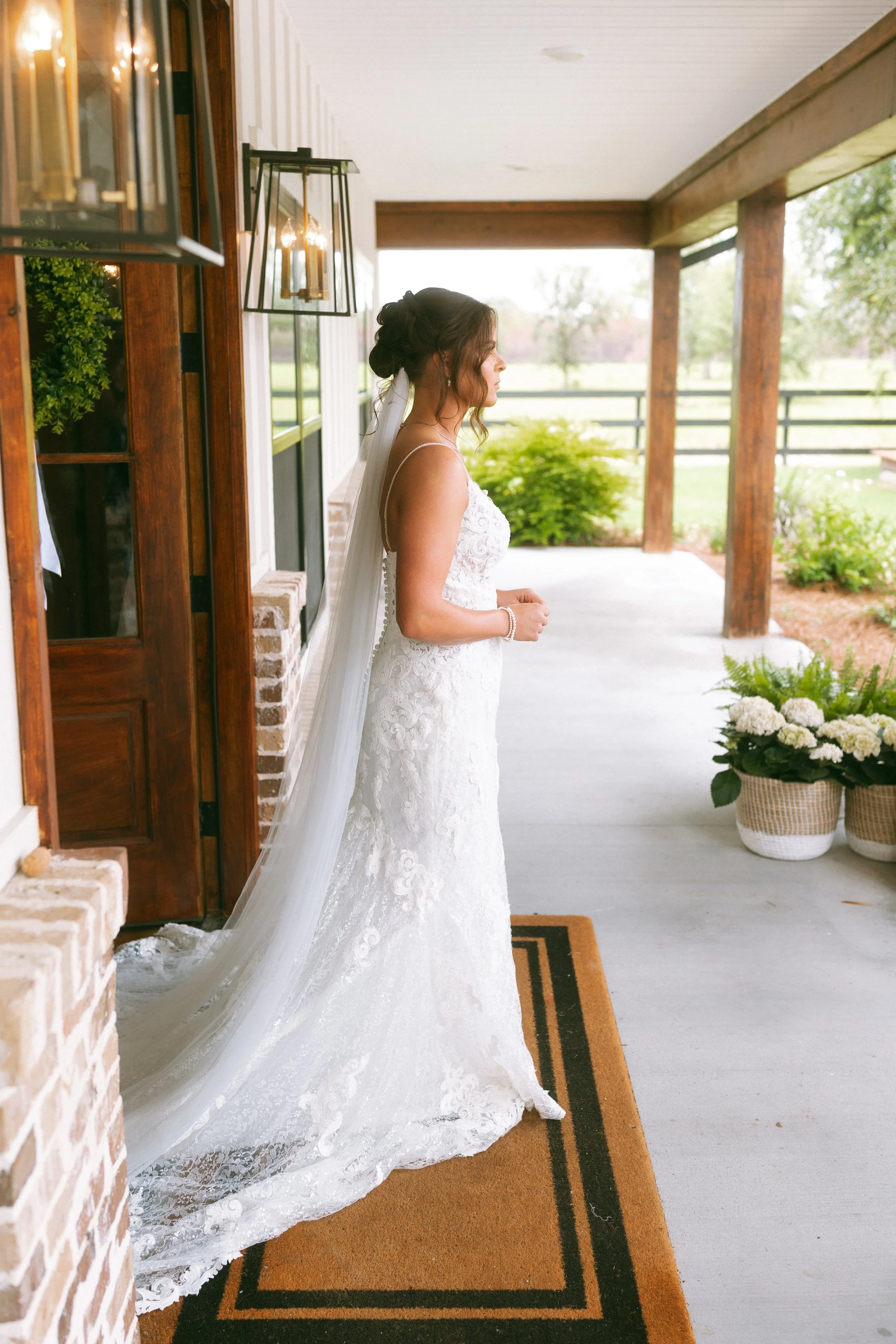 Emma stands on the covered porch at Red River Estate wearing a fitted lace wedding gown with a long veil and train.