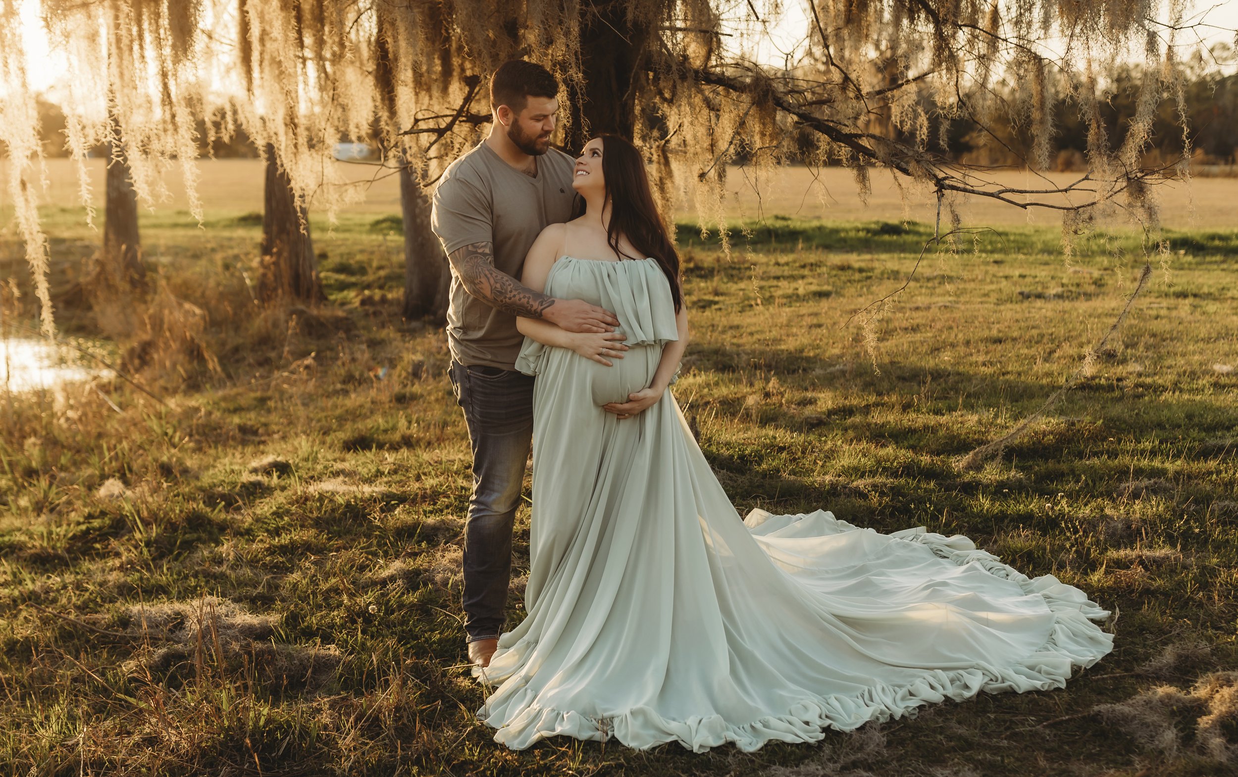 A couple standing outdoors under a tree with Spanish moss, the woman in a long white dress with a train, and the man in gray shirt and dark pants, embracing each other during sunset.