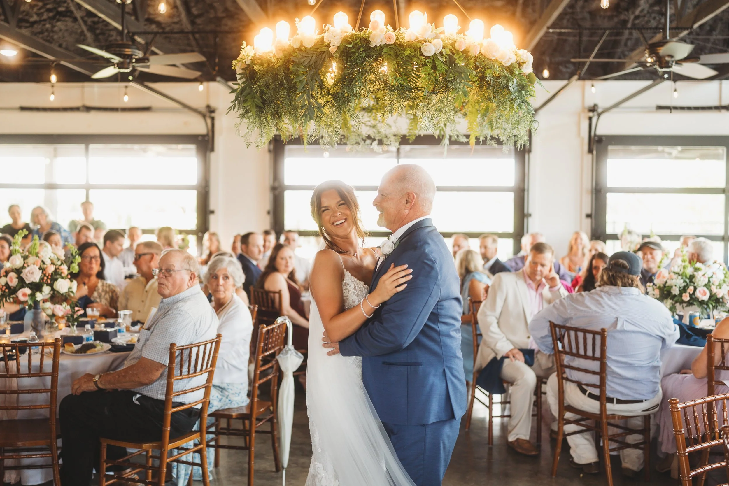 Emmie laughs while sharing a father-daughter dance beneath the floral chandelier during her reception at Red River Estate.