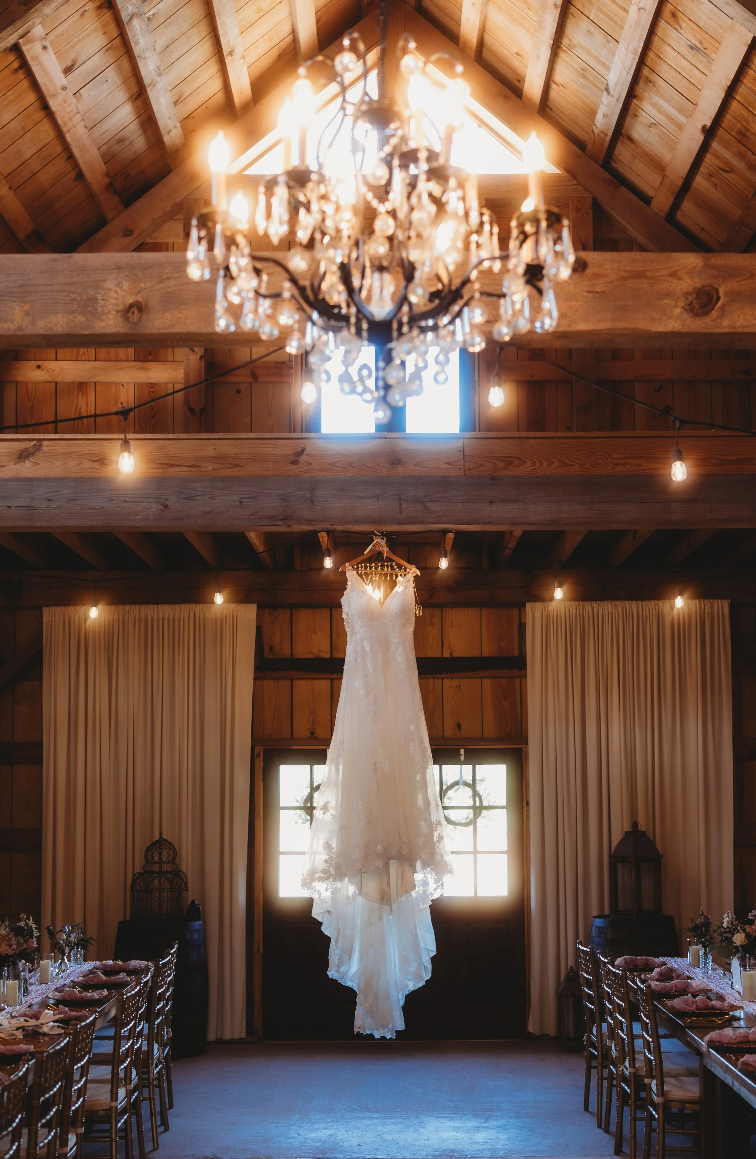 Bride’s wedding gown hanging in a softly lit barn beneath a glowing chandelier.