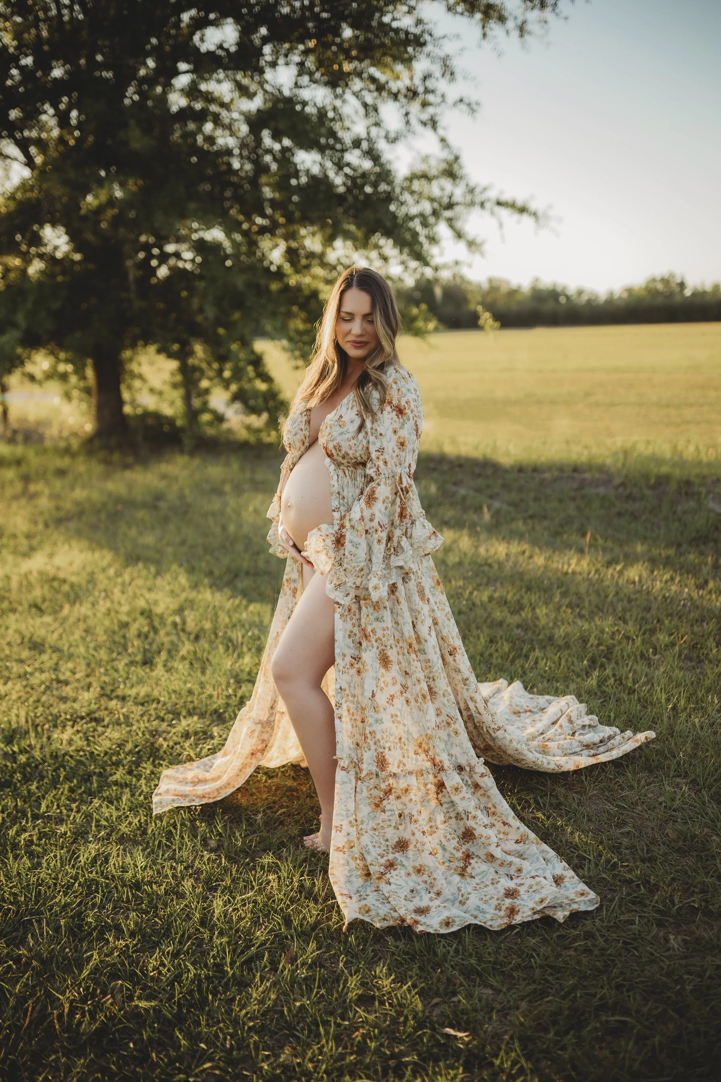 Golden hour maternity photo of an expecting mother in a flowing floral dress, standing barefoot in a peaceful field as she cradles her baby bump.