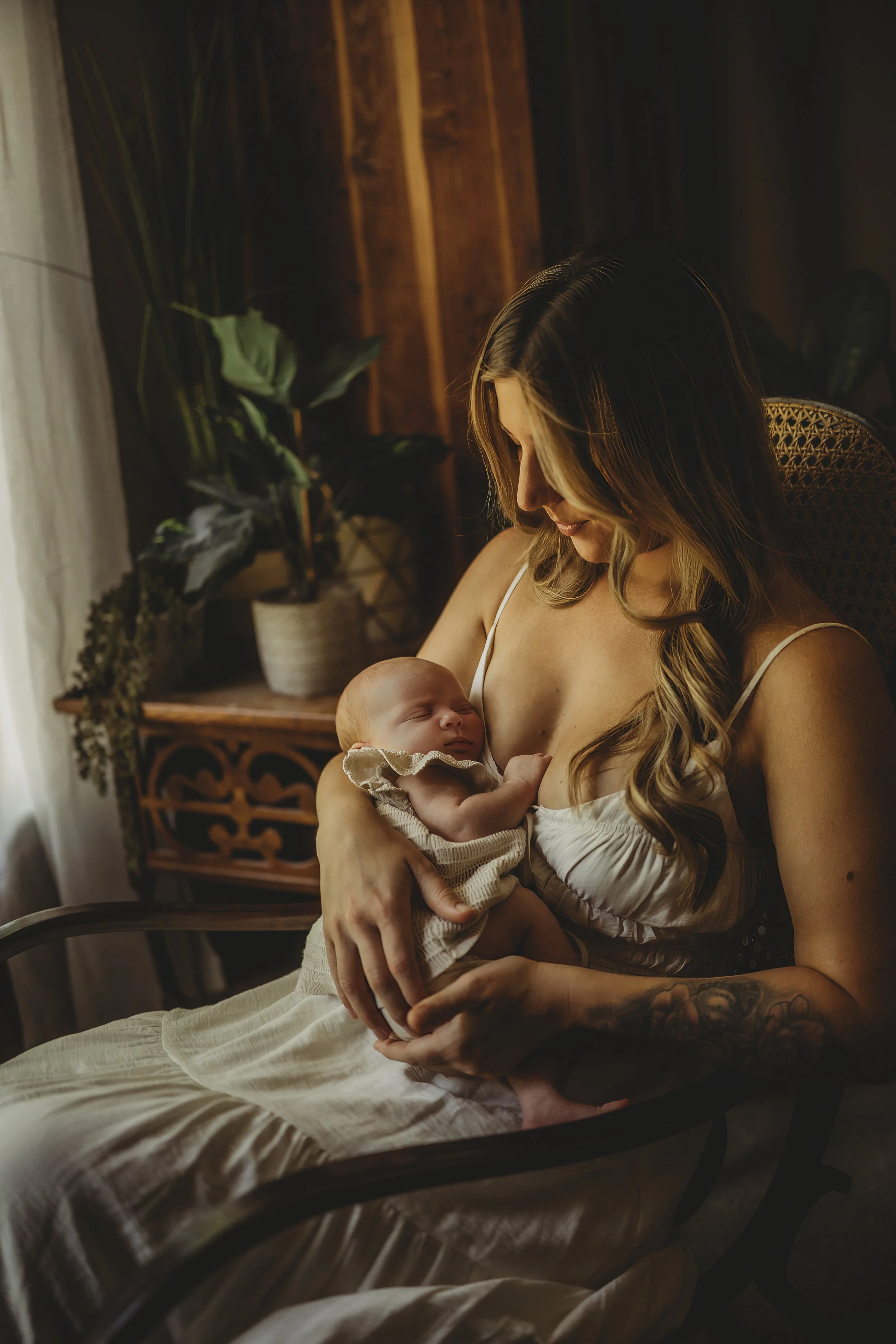 Mother seated in soft window light, cradling her sleeping newborn against her chest in a quiet, intimate moment at home.