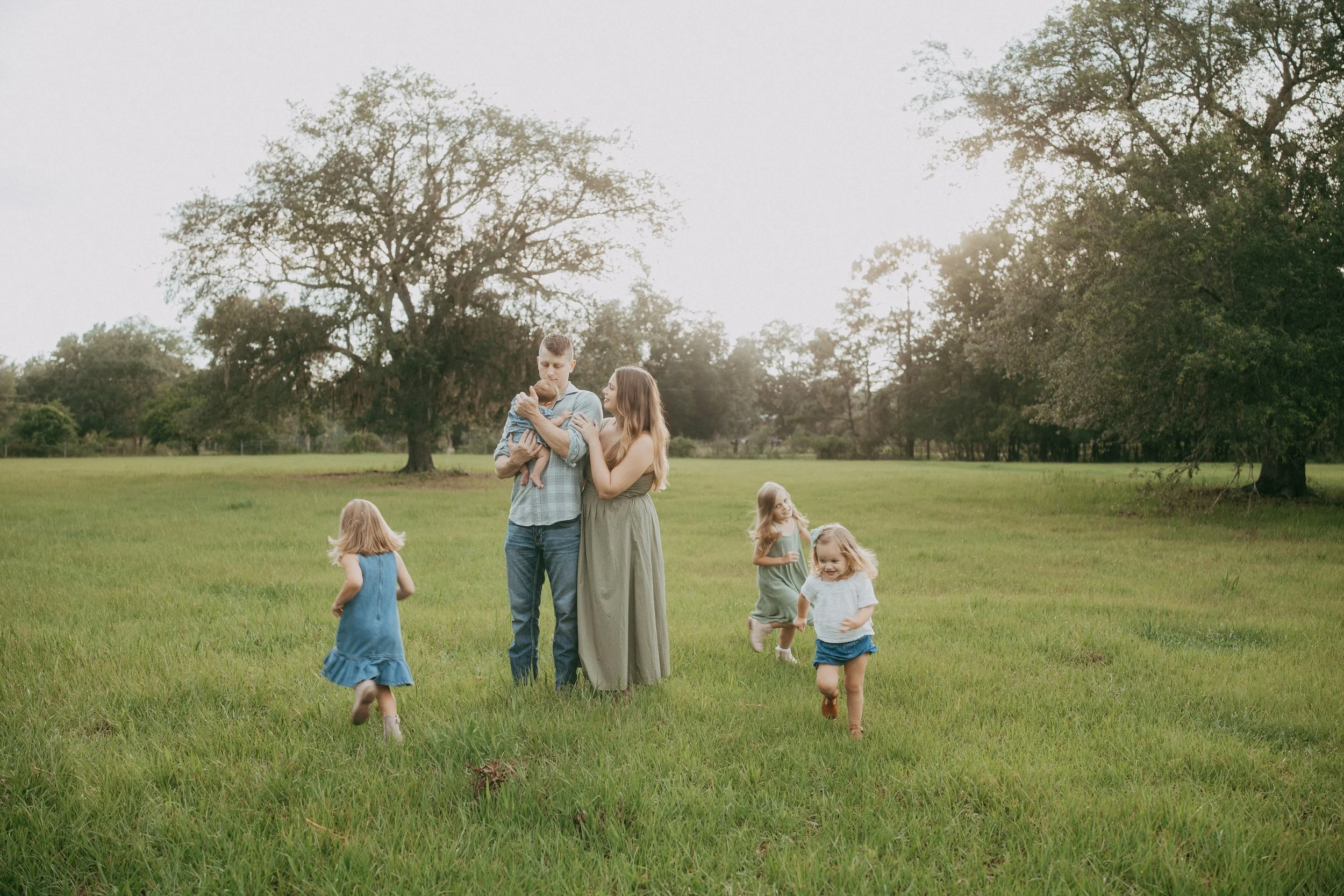 A family of six shares a playful outdoor moment in a sunlit field as the parents hold their baby and young children run happily around them.
