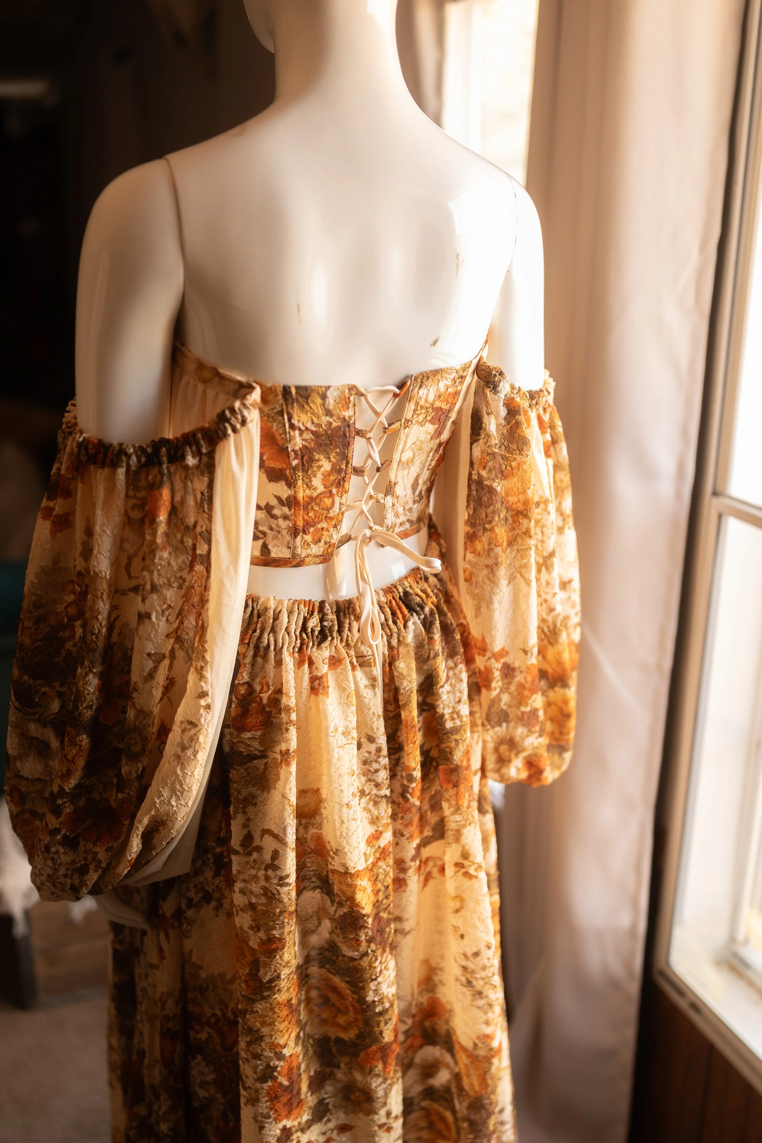Artistic floral maternity gown with lace-up back and soft draped fabric, styled indoors near a window.